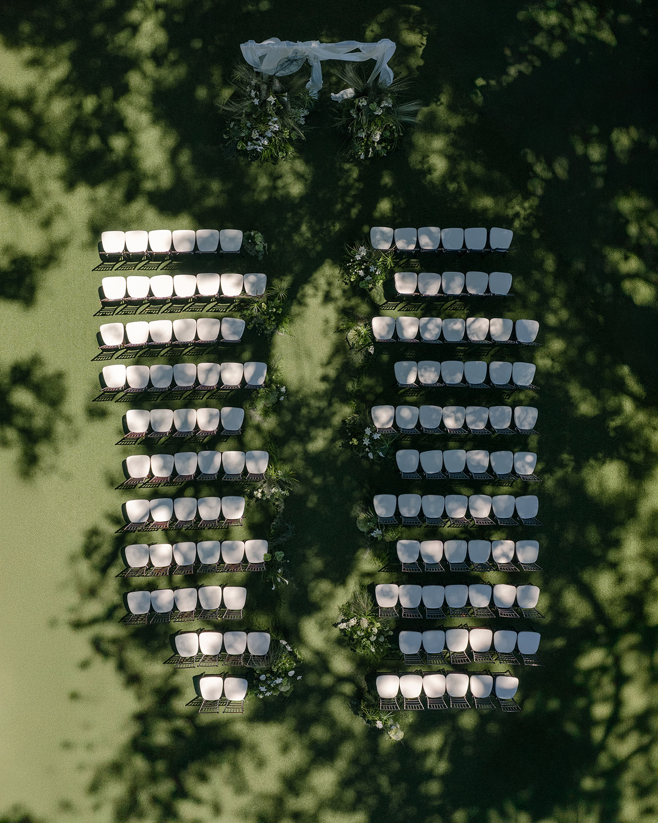 Aerial view of a beautifully arranged outdoor wedding ceremony at one of the best Oahu wedding venues, featuring rows of elegant white chairs, lush floral arrangements, and a soft white draped arch under the trees.