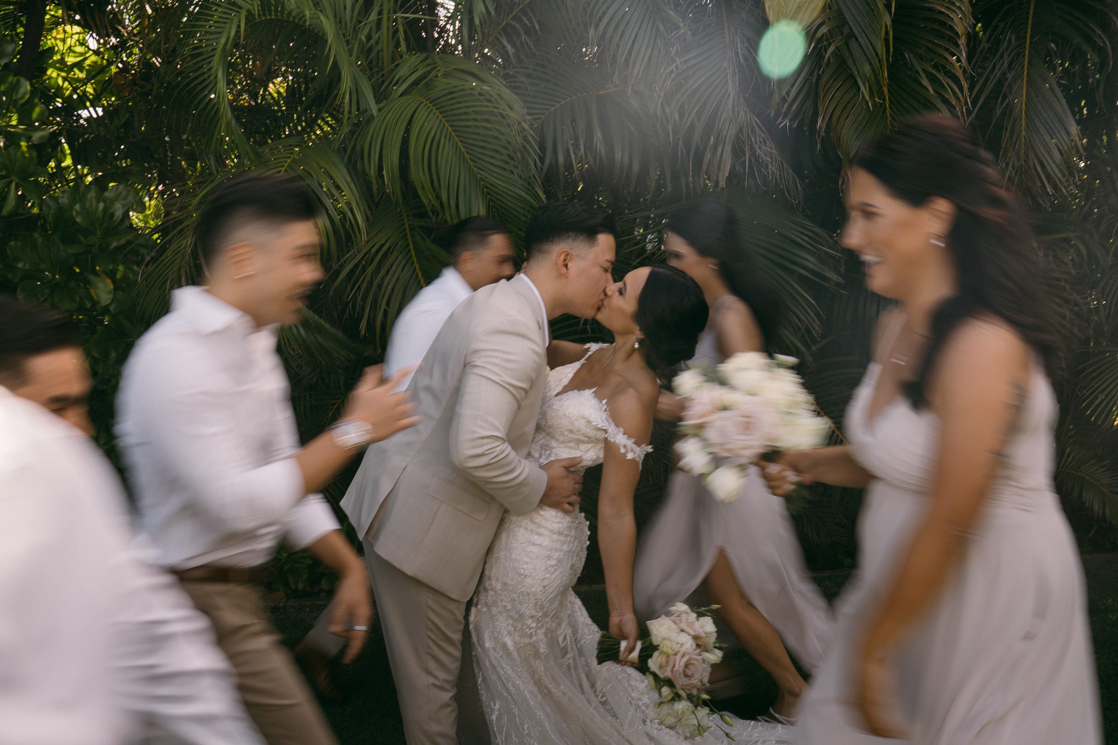 artistic portrait of bride and groom kissing while wedding party runs around them