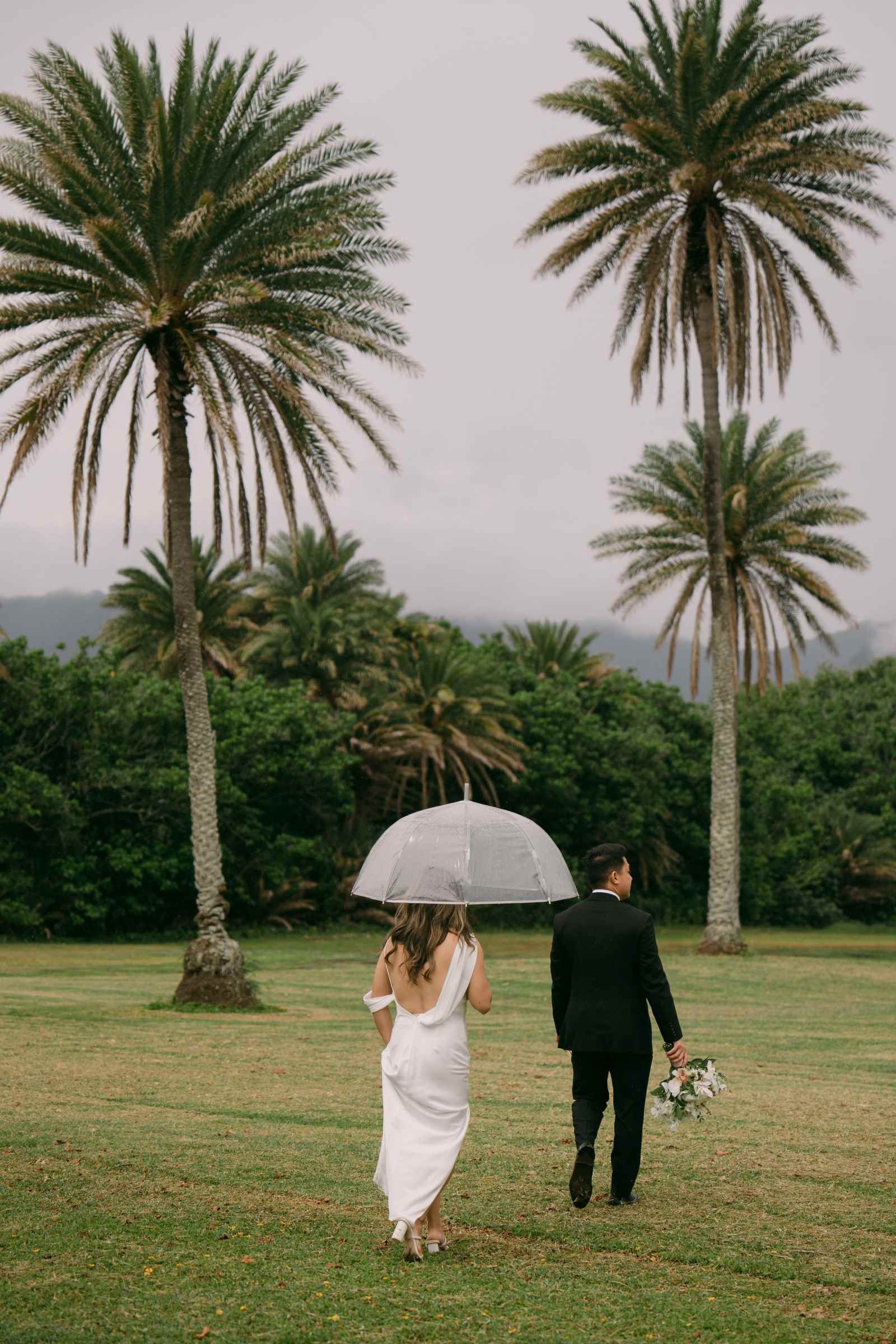 bride and groom walking with a clear umbrella on field in Hawaii