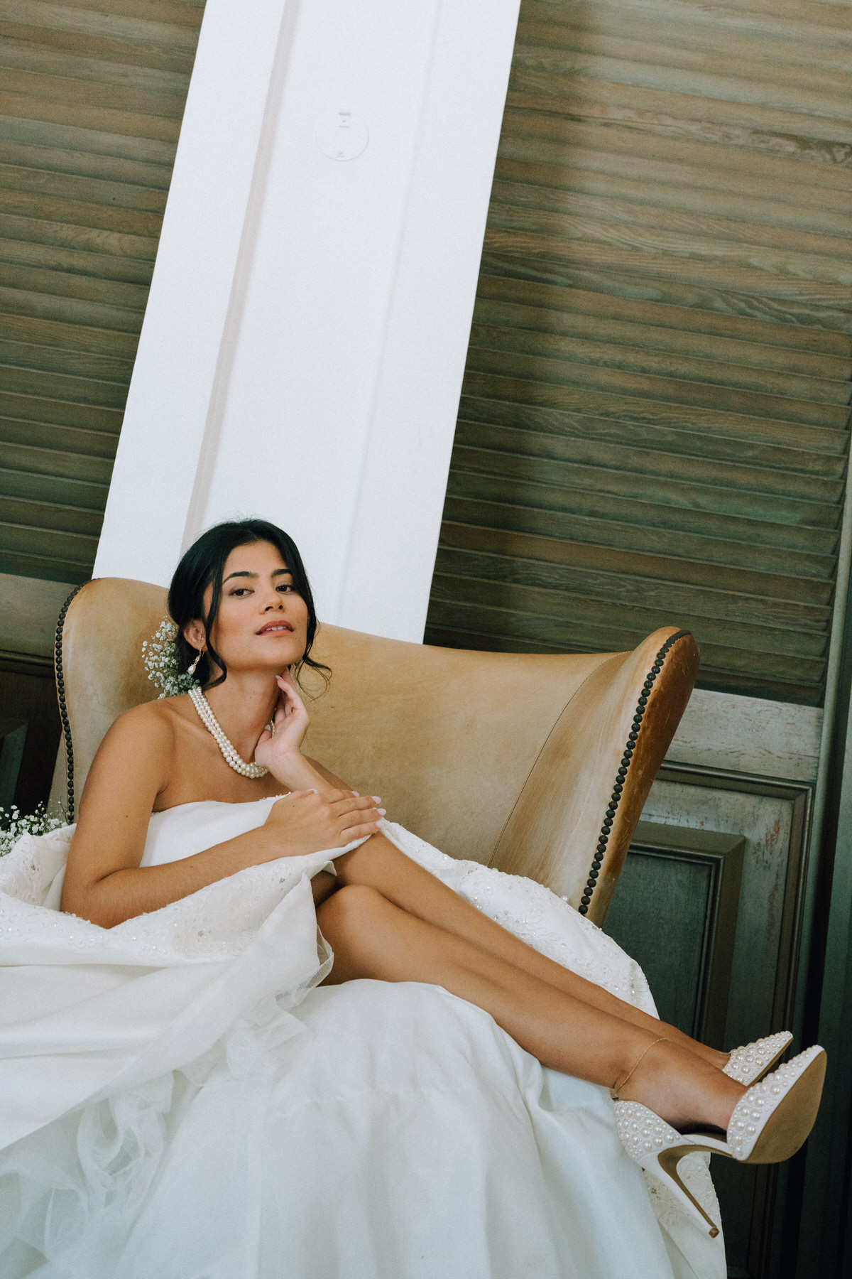 Bride sits in chair in contemporary wedding dress and shoes with pearls.