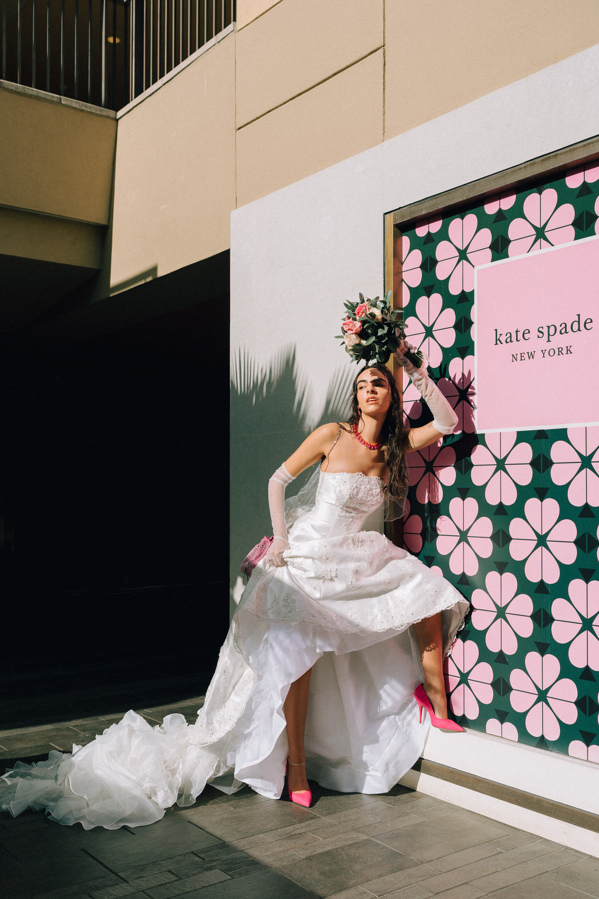 Bride poses in pink heels next to Kate Spade poster.