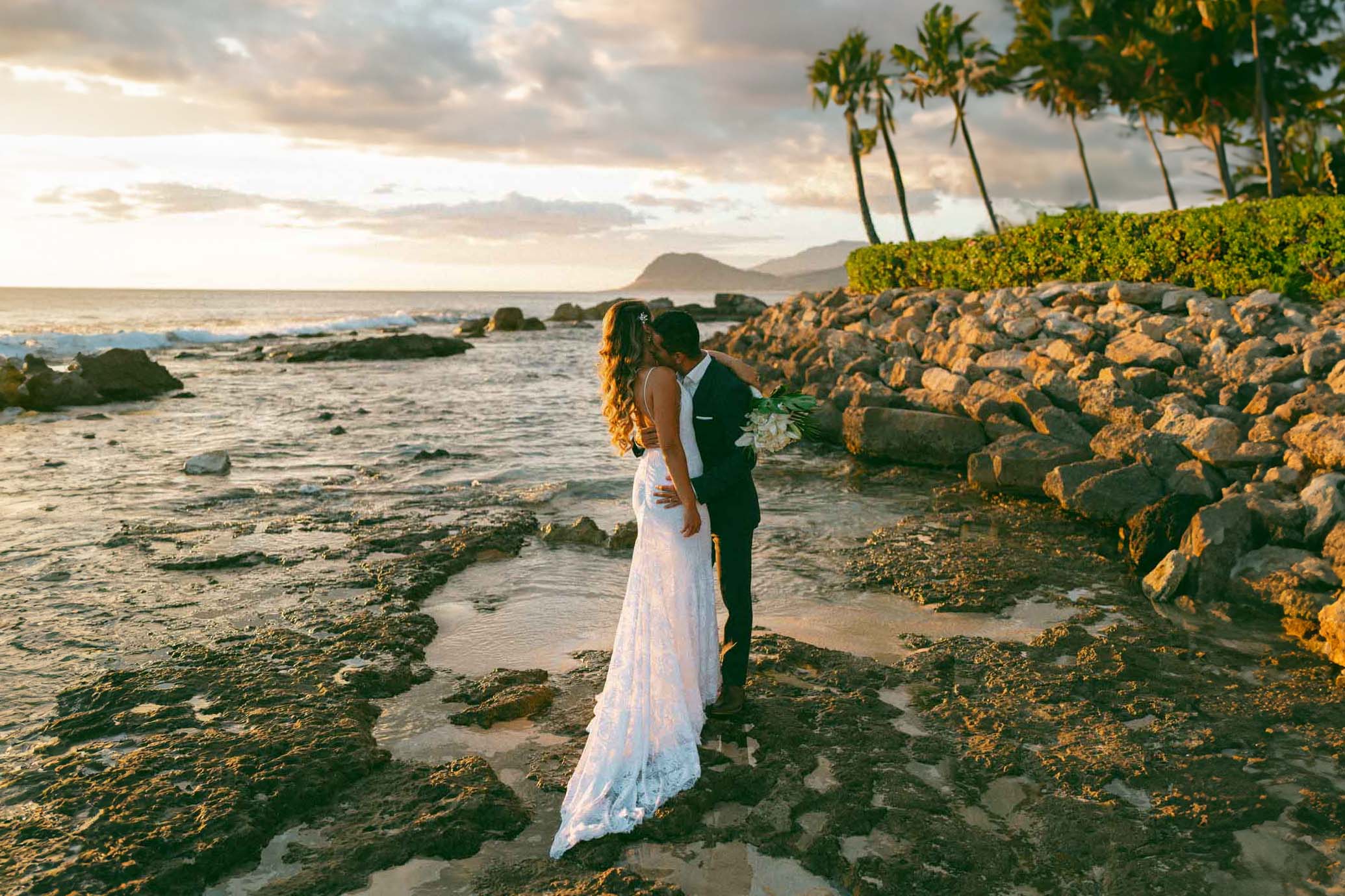 A close-up of a couple sharing a kiss, with a sunset backdrop and palm trees surrounding them.