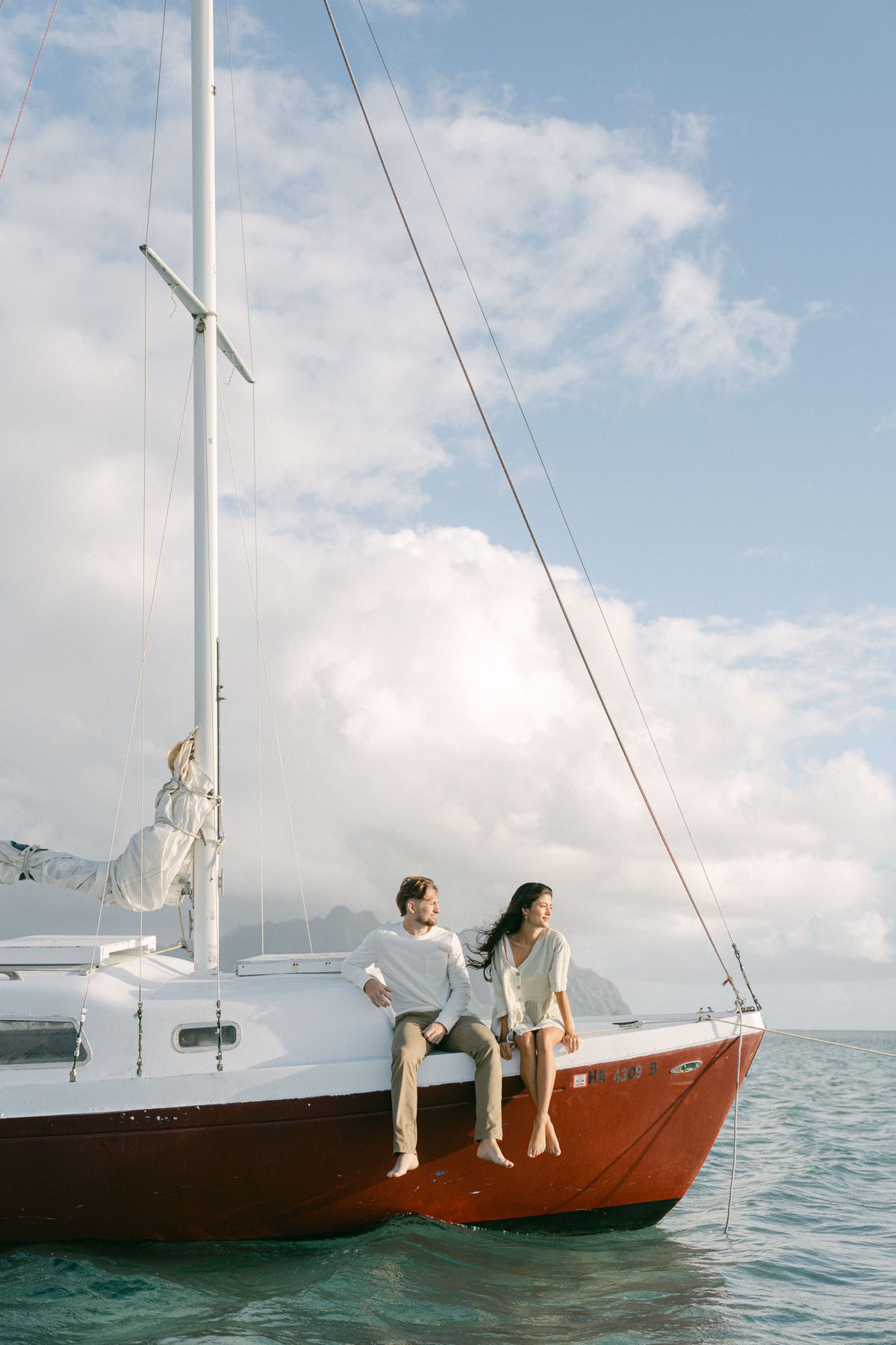 A joyful couple sits on the edge of a sailboat, looking out at the water. The woman smiles as the wind blows her hair, while the man sits beside her. The boat floats in clear waters with mountains visible in the distance under a partly cloudy sky captured by a wedding photographer in Waikiki.