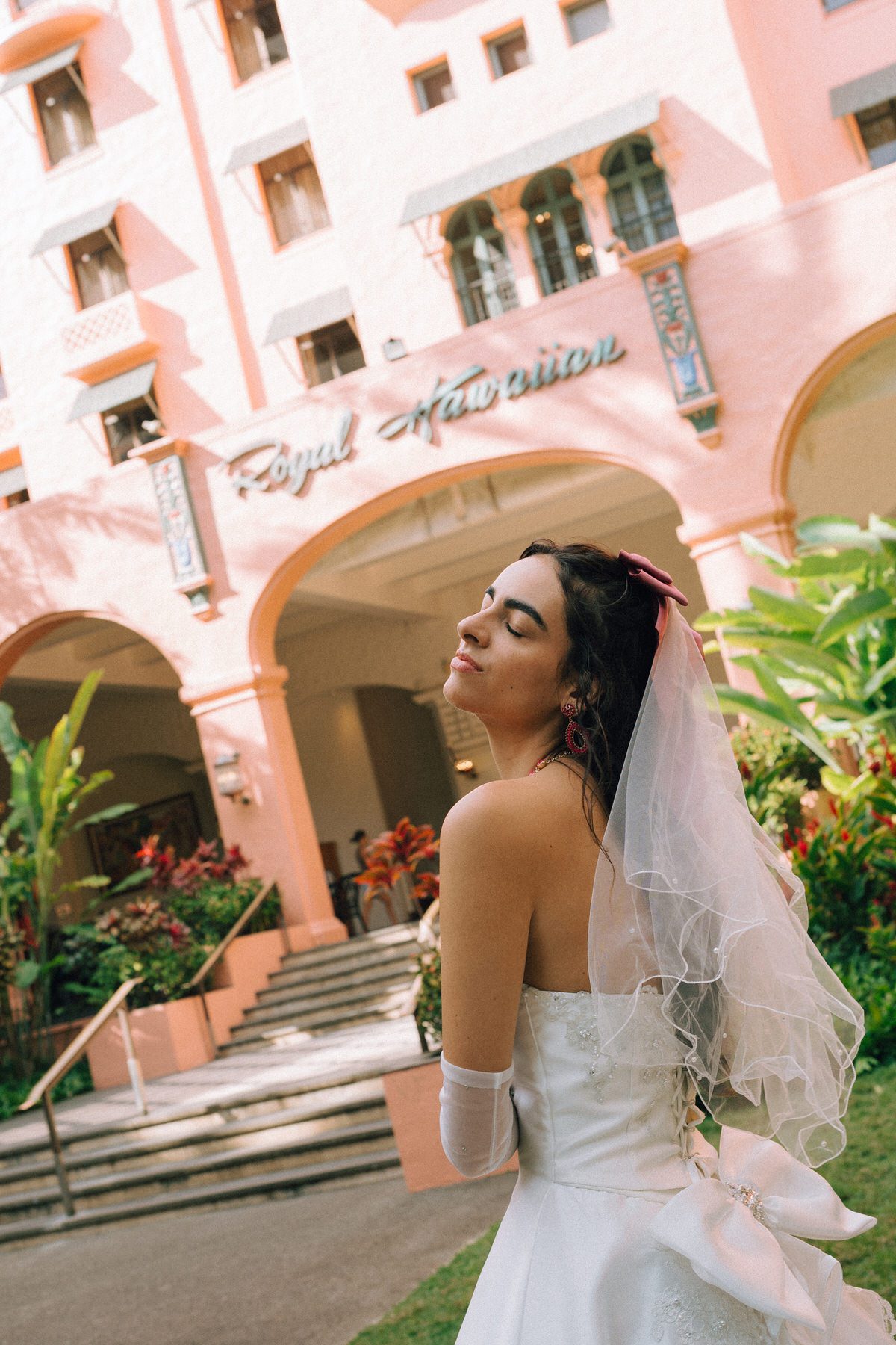 A bride standing in front of the Royal Hawaiian Hotel, her veil catching the sunlight as she tilts her head back in a carefree moment.