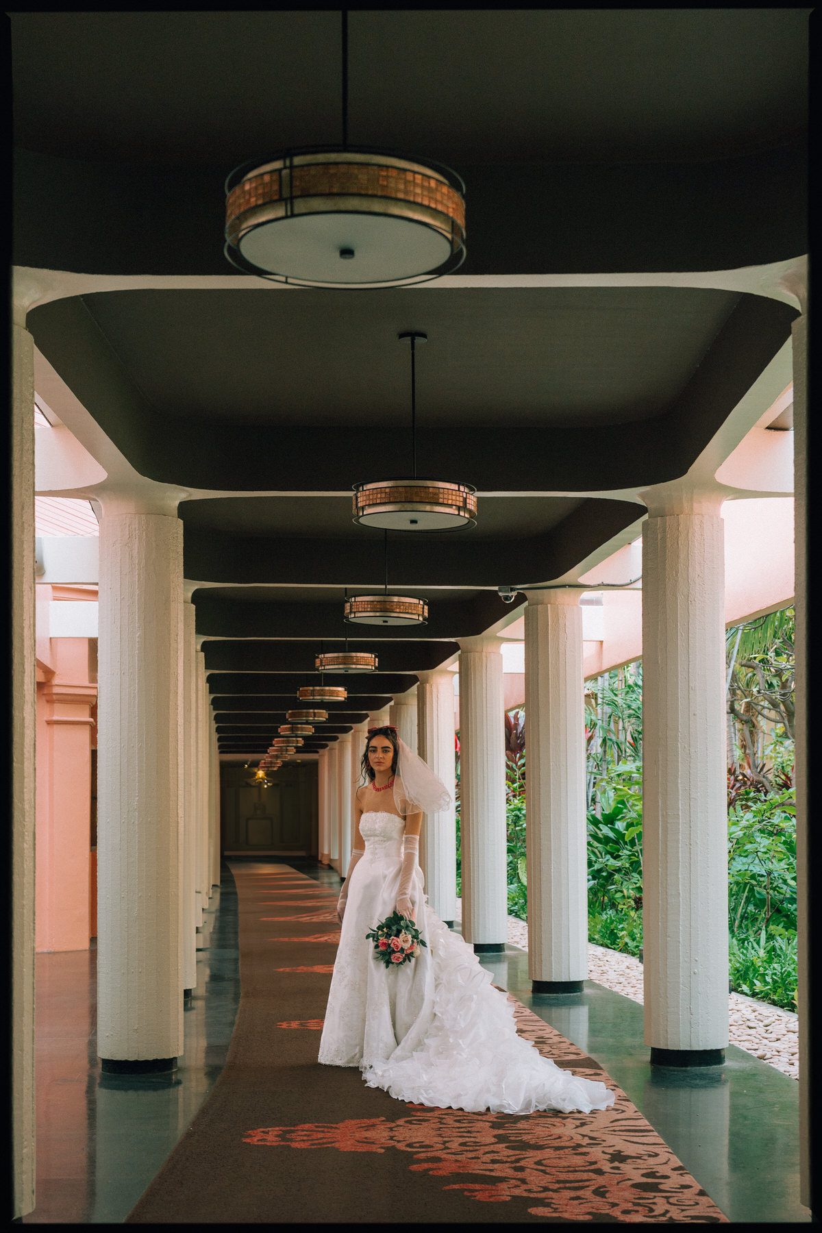 A bride walking down a grand hotel corridor, carrying a bouquet, with soft light filtering through the pillars.