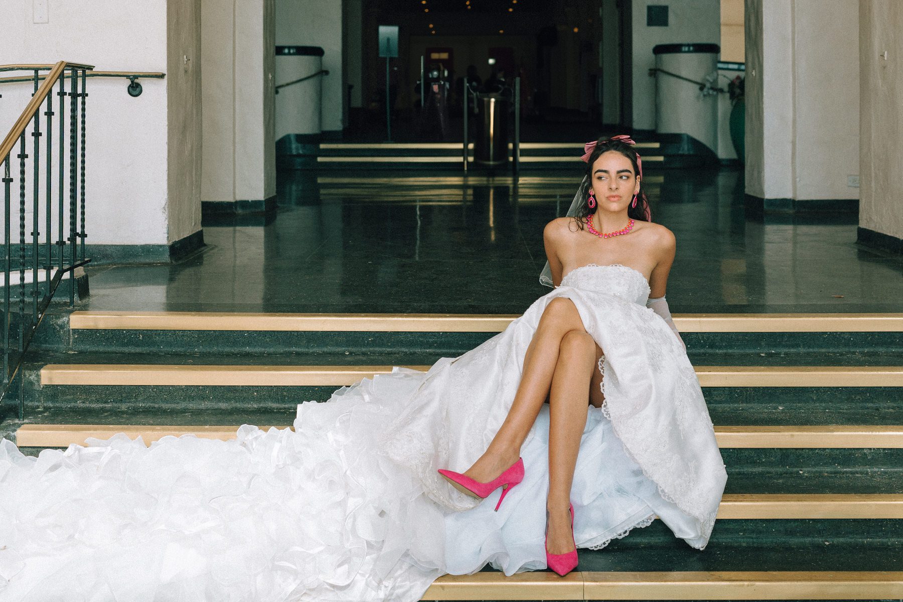 A second version of the bride sitting on the steps, showcasing her dramatic gown, vibrant pink accessories, and effortlessly chic attitude.