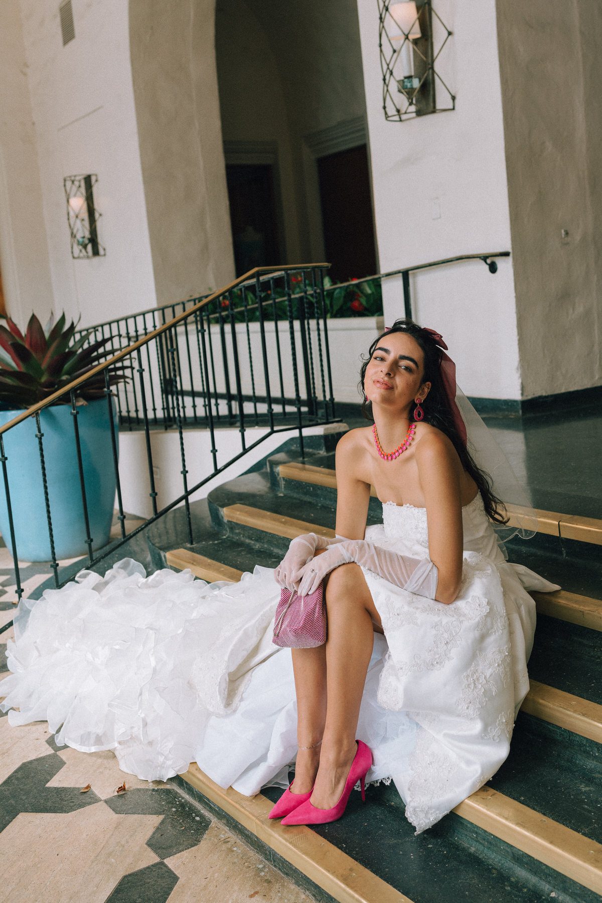 A stylish bride sitting on the steps of a historic hotel, wearing hot pink heels, gloves, and a matching handbag, adding a playful twist to her classic wedding dress.