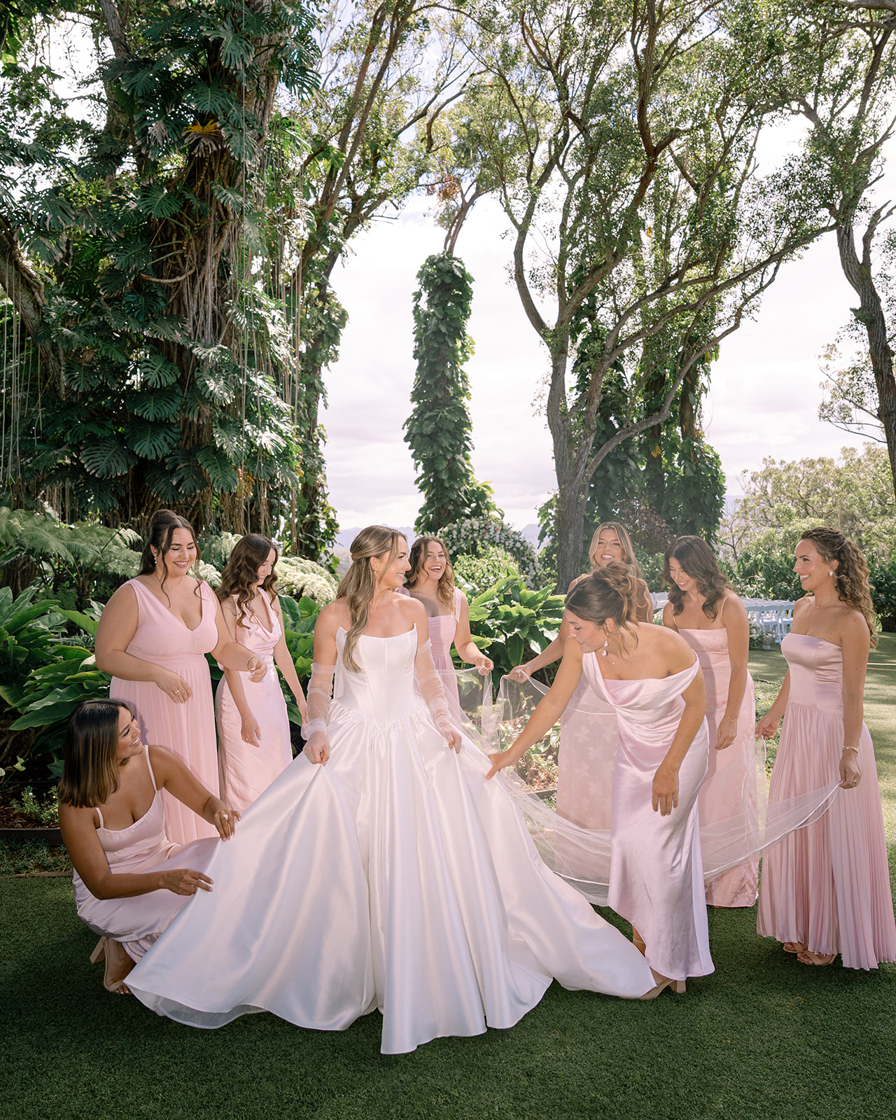 Bride surrounded by bridesmaids in blush pink dresses, adjusting her gown in a lush garden setting at Sunset Ranch Oahu — a popular choice among luxury Hawaii wedding venues.