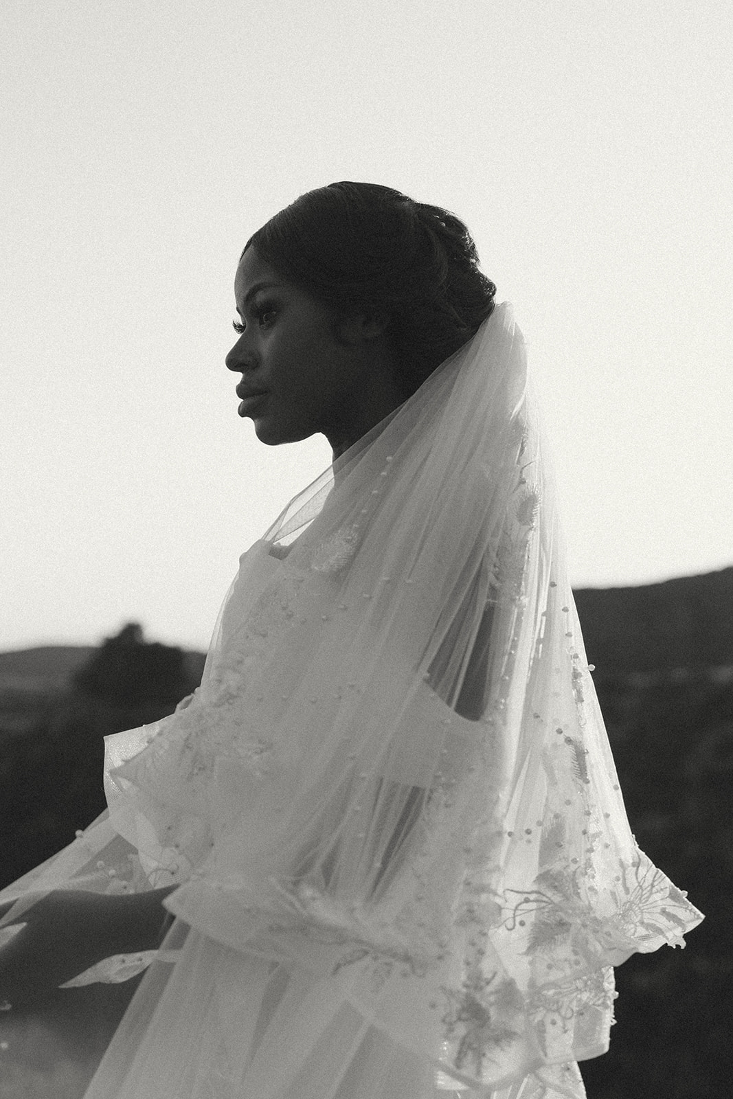 A black and white portrait of a bride in a delicate lace veil, gazing into the distance with a serene expression.