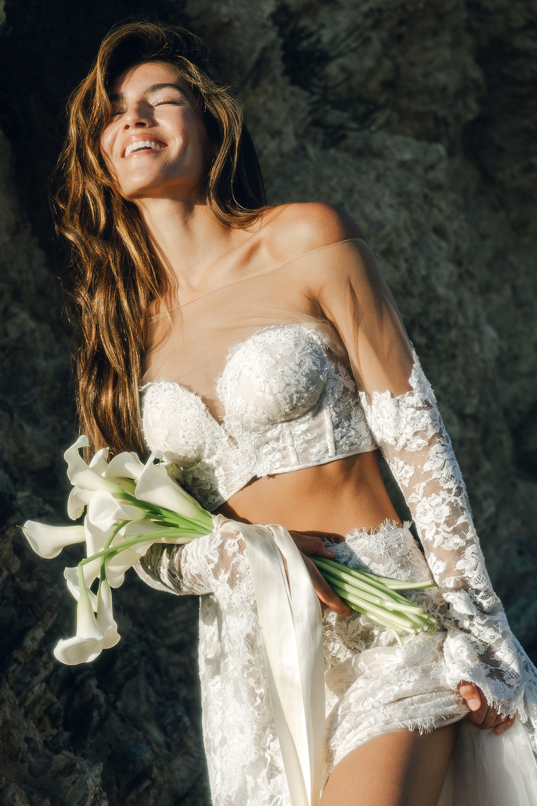  A bride laughing effortlessly by the rocks, holding calla lilies, wearing a chic lace corset and sheer sleeves.