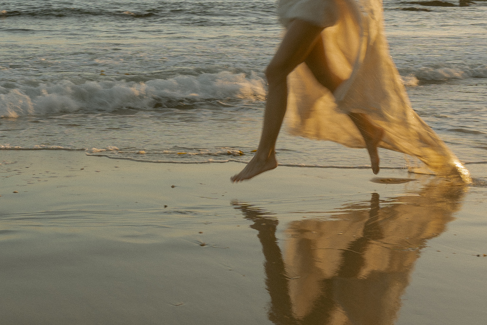A dreamy, golden-hour shot of a bride running barefoot along the shoreline, her dress flowing in the wind and reflecting in the wet sand.
