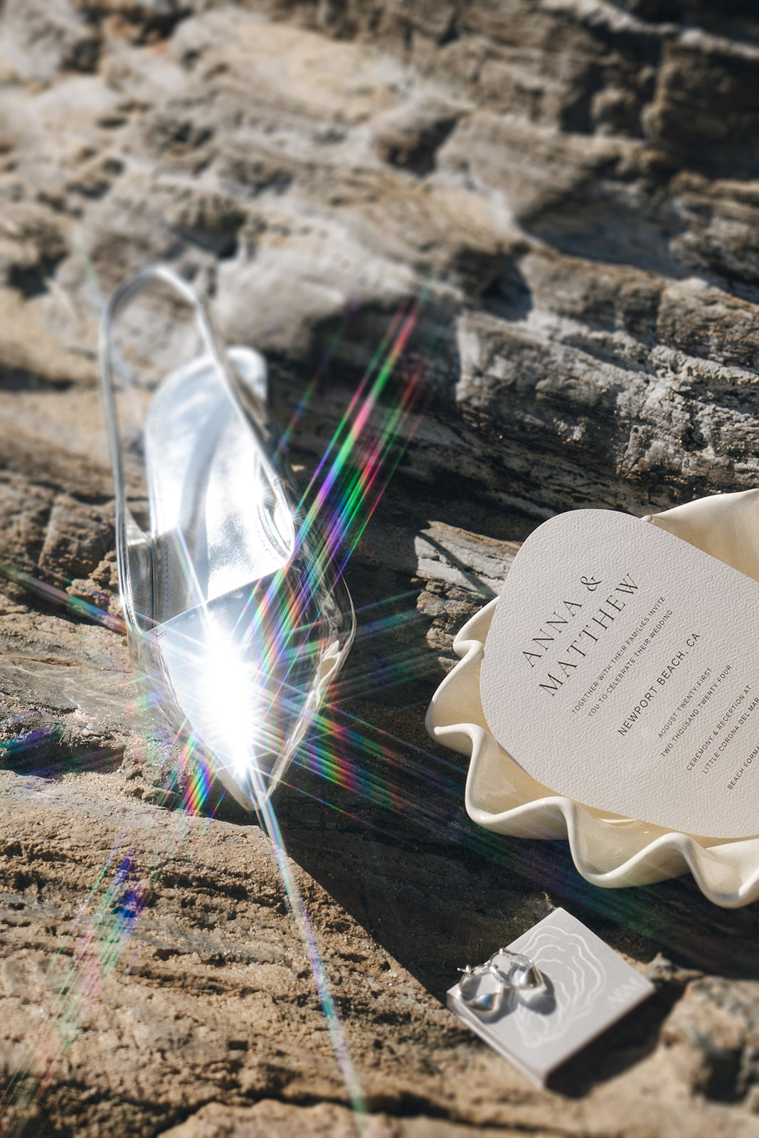A wedding flat lay featuring a reflective silver shoe, a modern invitation card, and pearl earrings placed on a textured rock surface with a sun flare.