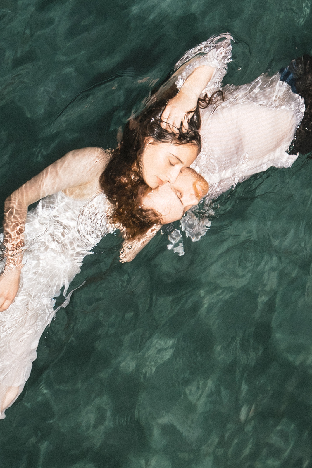 A couple is floating in the water, embracing each other. The woman is wearing a white dress, and the man has a light-colored s
