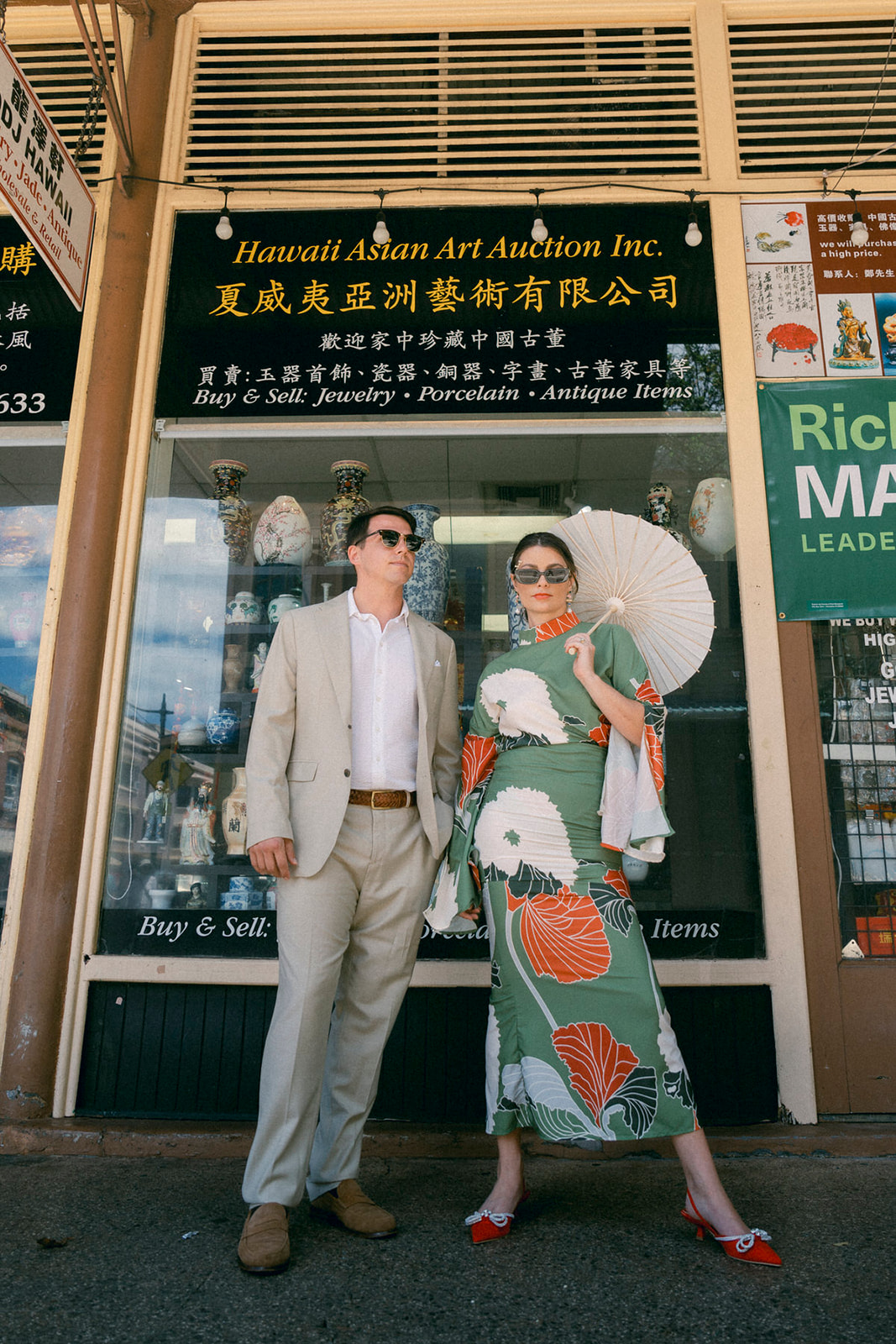 A couple stands in front of a store with a sign that reads "Hawaii Asian Art Auction Inc." The woman is wearing a green dress with large floral prints, holding a white parasol, while the man is dressed in a beige suit and sunglasses.