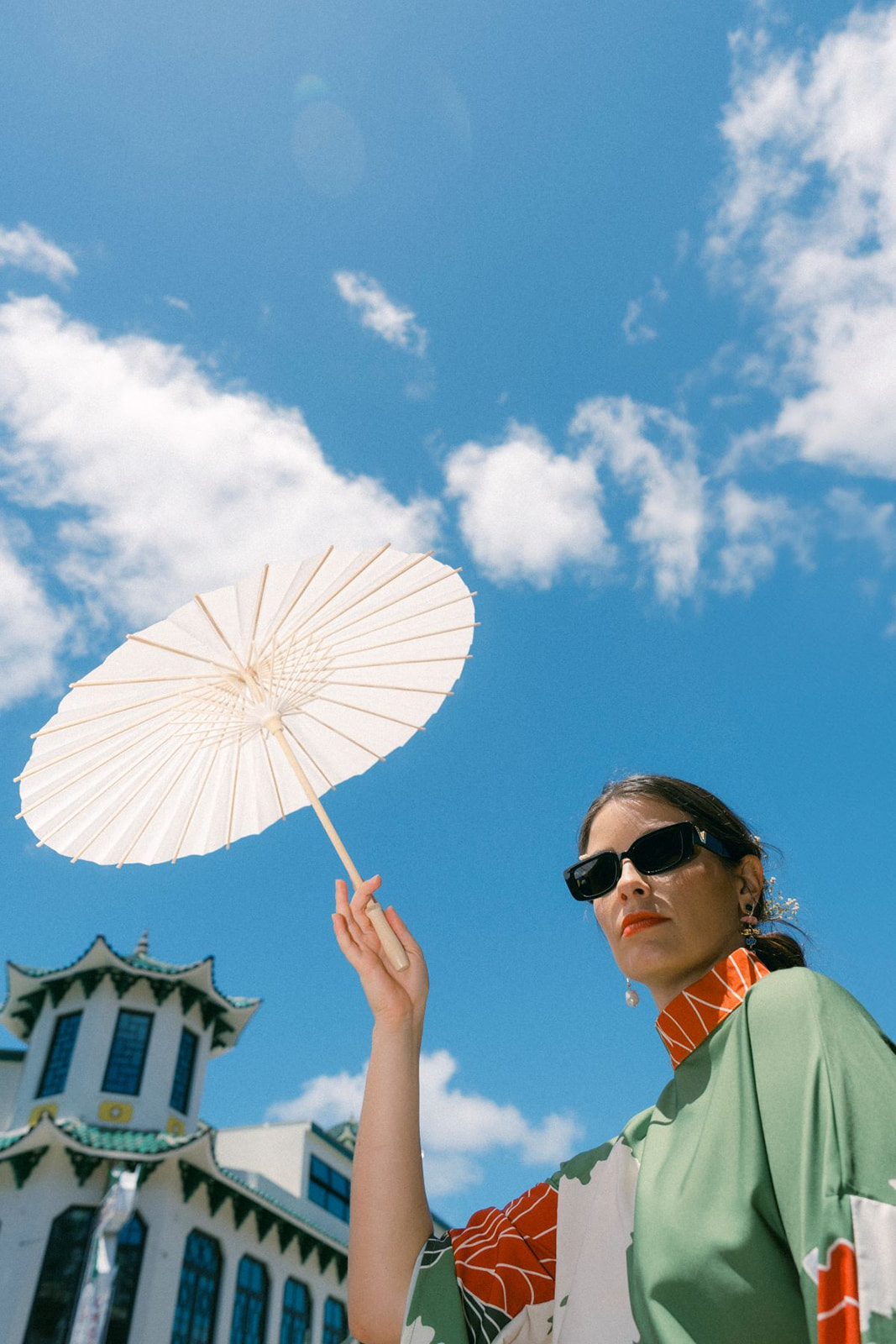 A woman holding a white parasol against a bright blue sky, wearing sunglasses and a green patterned dress, with a Chinese-style building in the background.