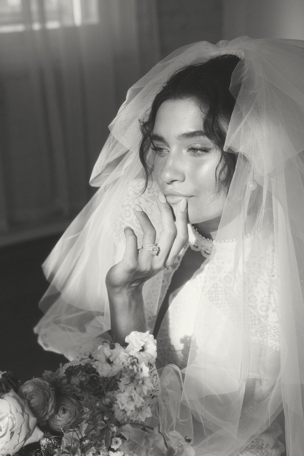 A close-up of a bride resting her hand on her lips, soft light and shadows highlighting the delicate veil and floral bouquet.