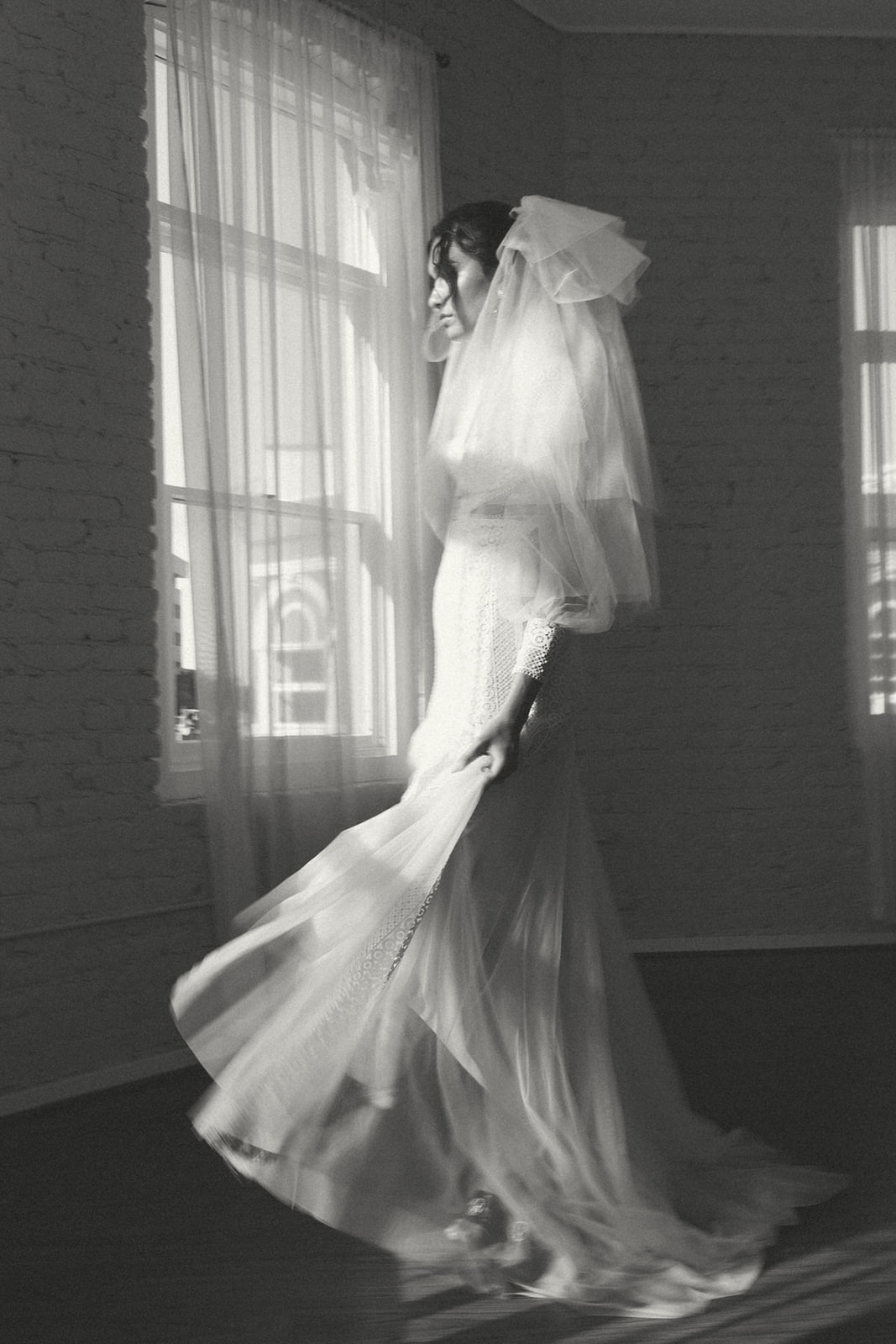 A bride in a flowing lace gown and veil, mid-twirl, caught in soft window light with an elegant, timeless feel.