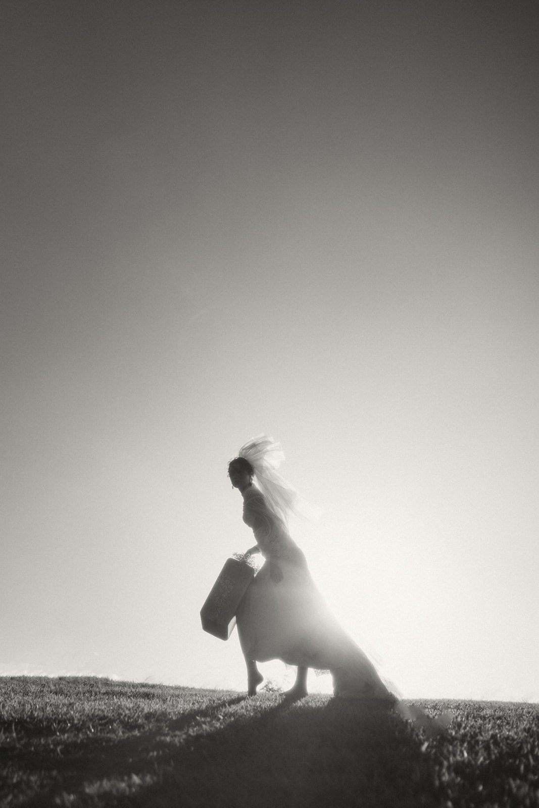 A black and white silhouette of a bride walking up a hill at sunset, carrying her flowing dress and a woven bag.
