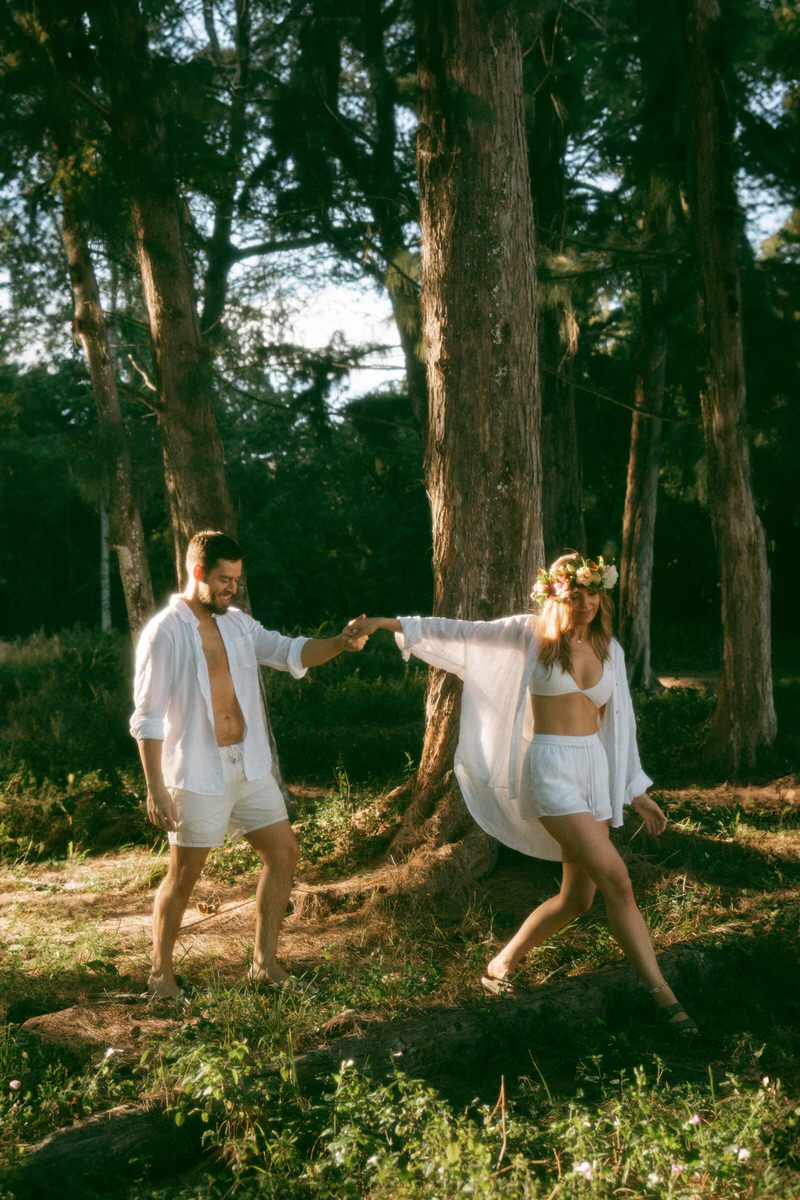 A couple walks through a forested area, dressed in white, with the woman wearing a floral crown and the man shirtless, creating a romantic vibe against the backdrop of tall trees. Their carefree stroll captures the beauty of their connection, ideal for a romantic photography session in Kaneohe.
