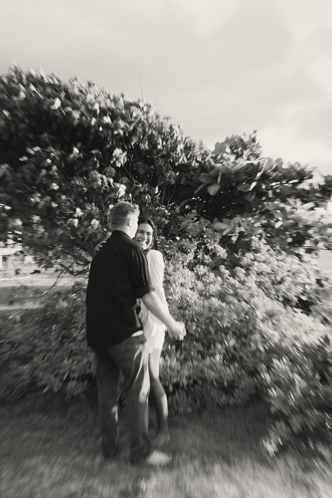 A black-and-white photograph showing a couple playfully spinning around in front of a lush, leafy tree. The image has a dreamy, blurred effect, capturing the joy of the moment.