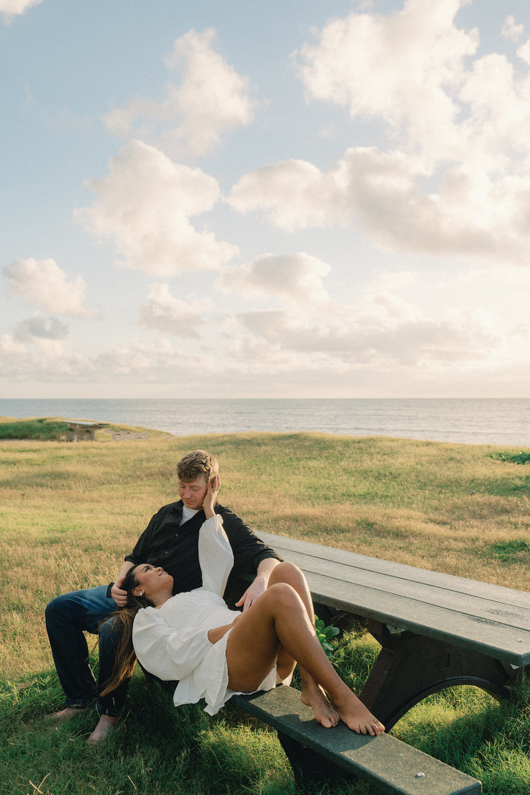 A couple sitting on a park bench near the ocean, with the woman reclining and playfully resting on the man's lap, bathed in warm sunset light.