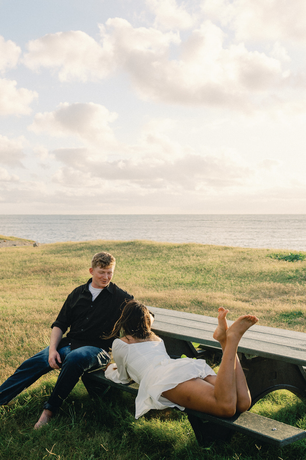 A playful couple enjoying each other's company, the woman lying on the bench with her feet up, while the man sits casually next to her, against a peaceful coastal backdrop.
