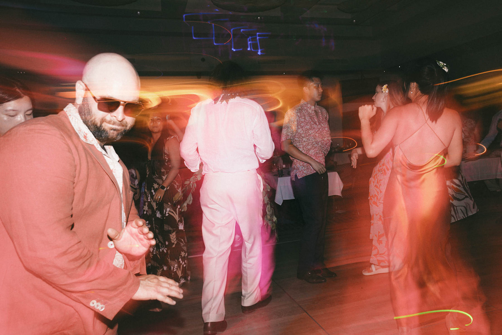 A lively dance scene at a party with a mix of people dancing. A bald man in a red jacket with sunglasses is in the foreground, while others in colorful outfits dance in the background, with streaks of vibrant light creating a dynamic atmosphere.