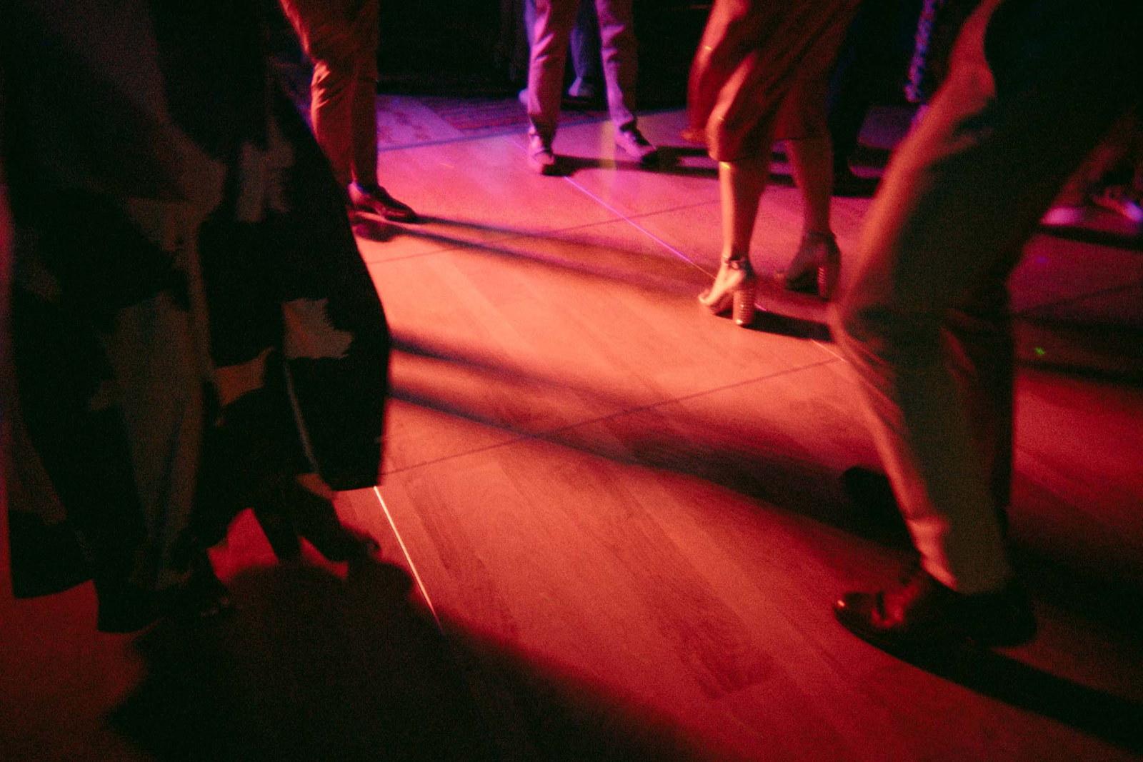 A group of people dancing on a wooden floor, their feet in motion. The image captures a vibrant atmosphere with red lighting reflecting off the floor and shadows of the dancers' legs.