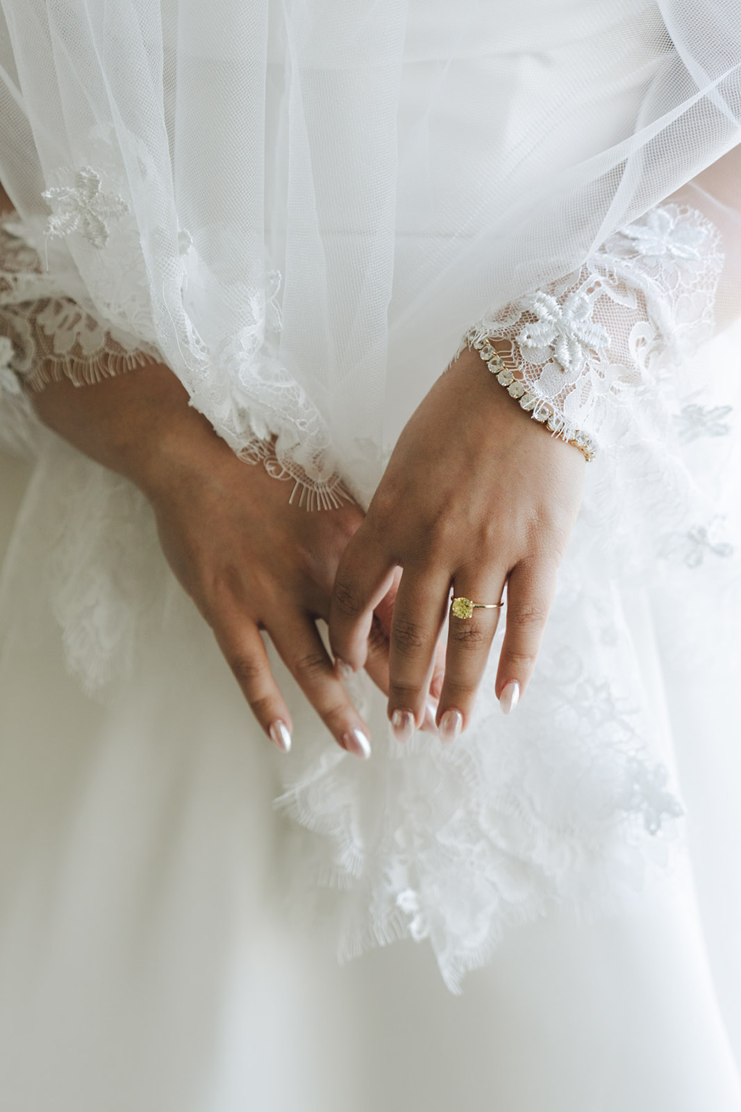 A close-up of a bride’s hands gently folded over her dress, adorned with a delicate lace veil, gold ring, and beaded bracelet.