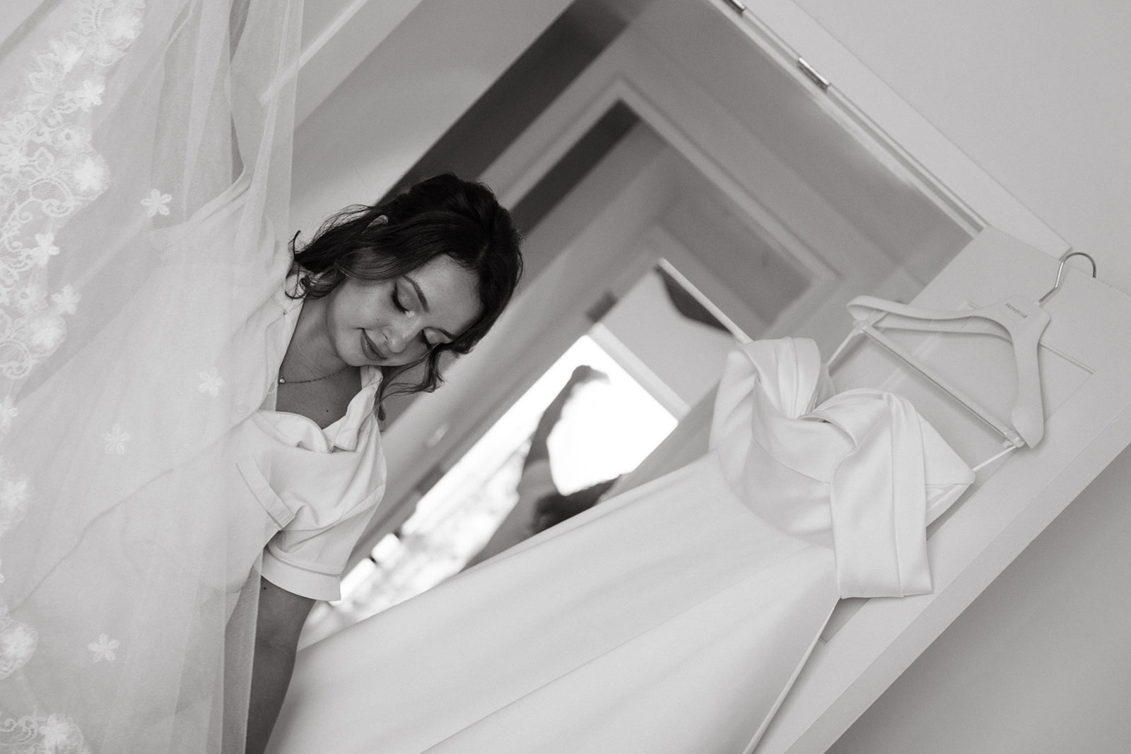 A bride, smiling softly while holding her veil and dress, with her wedding gown hanging in the background.