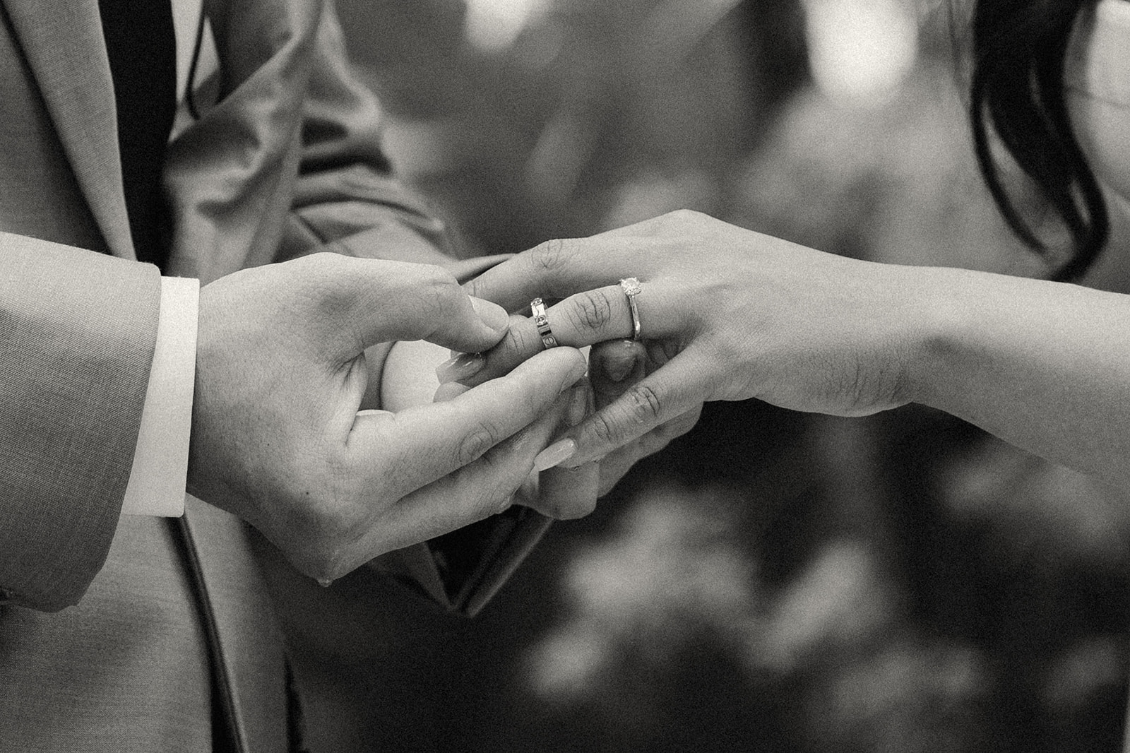 A close-up black and white image of two hands exchanging wedding rings.