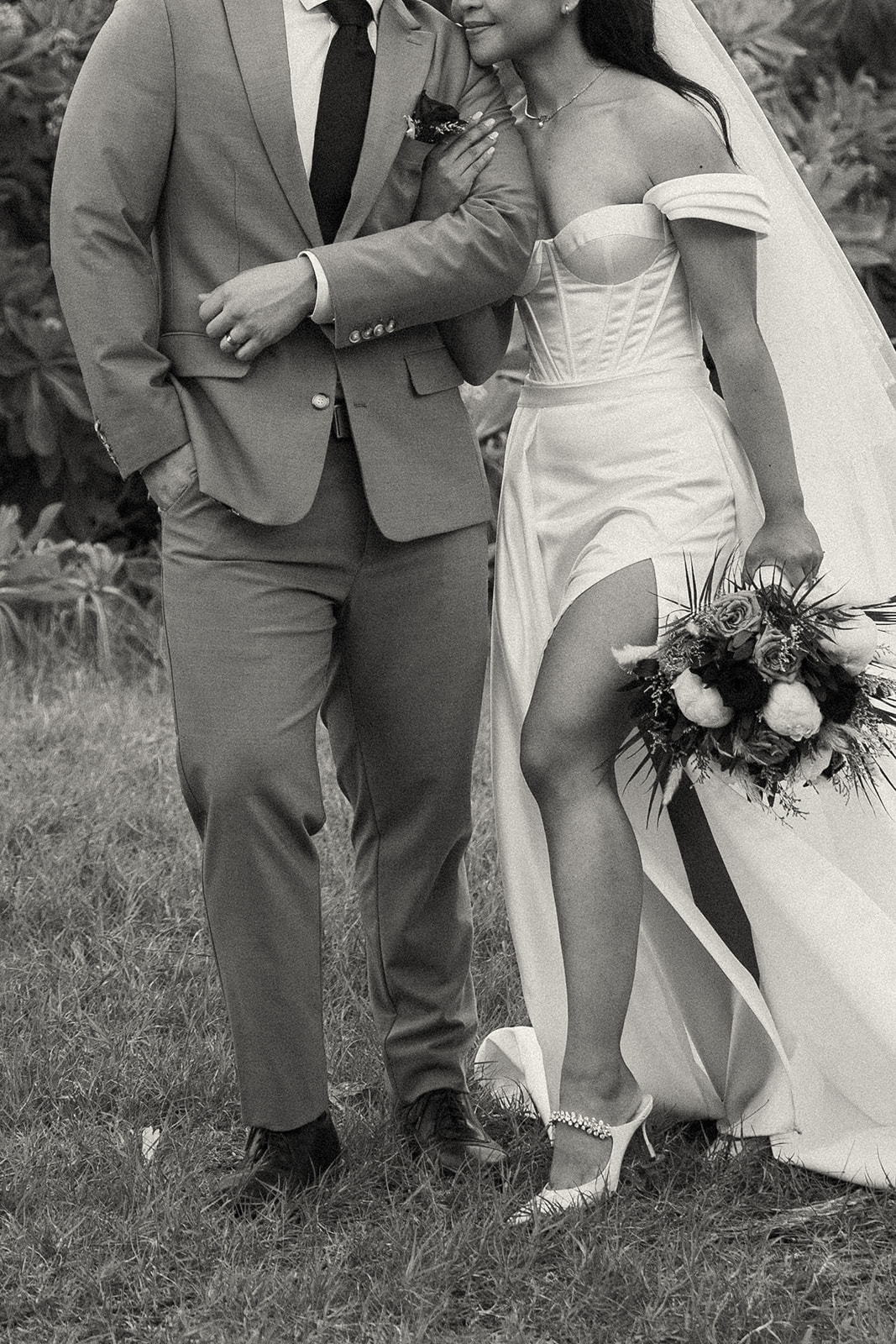A bride and groom standing together, with the bride holding a bouquet and wearing a corset-style wedding dress, revealing her wedding heels.