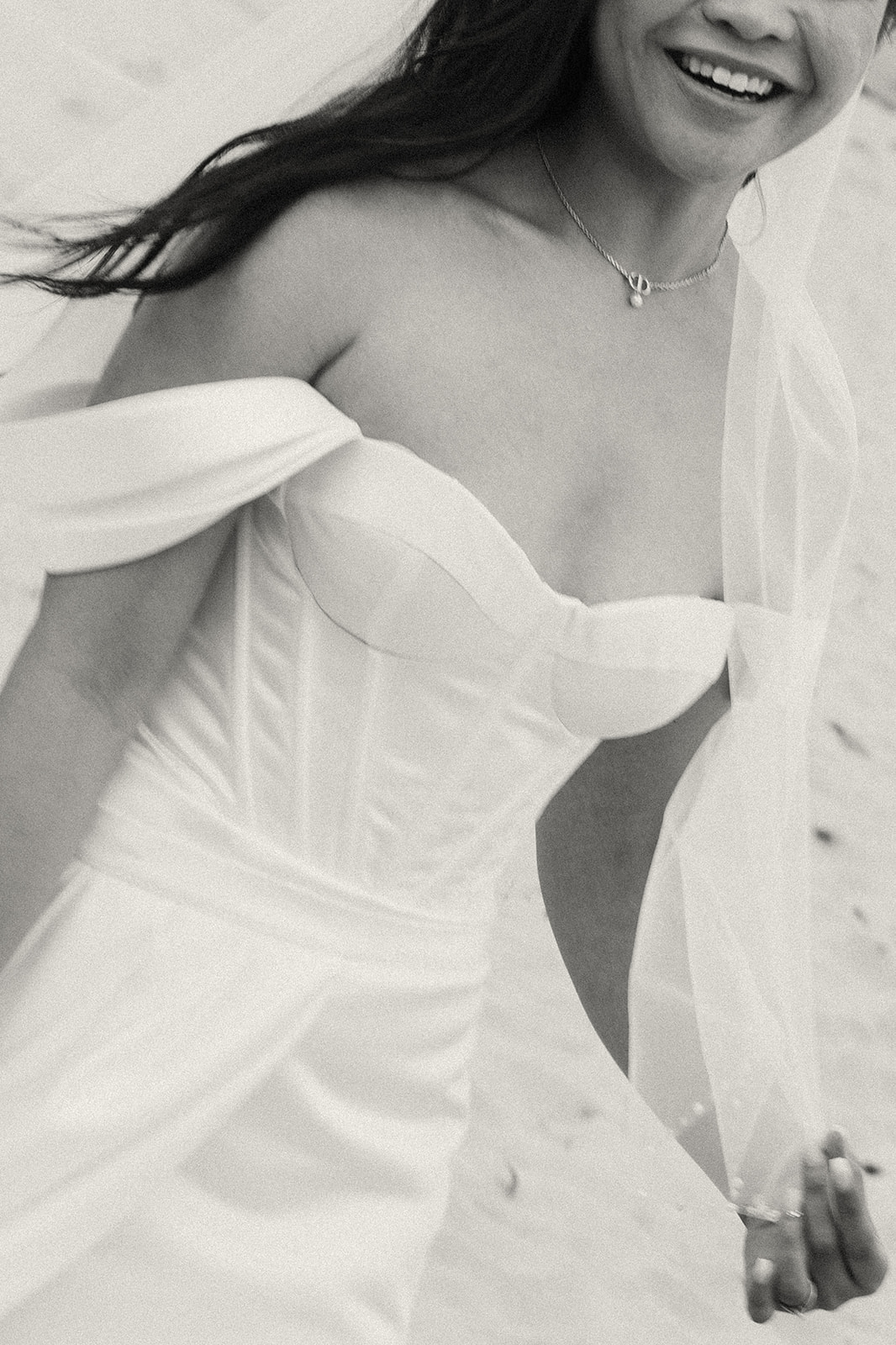 A candid black and white image of a smiling bride in her corset-style wedding dress, with her veil flowing as she walks.