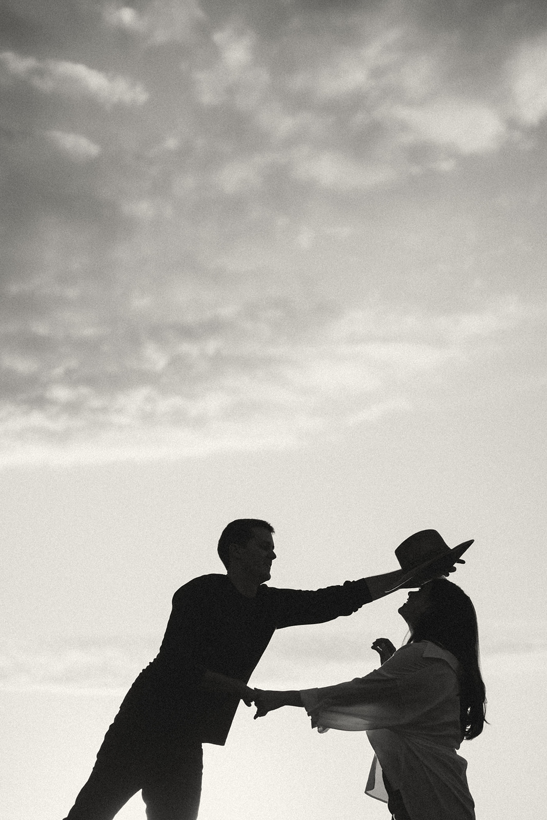 A silhouette of a couple dancing on a beach at sunset. The man is playfully holding the woman's hat while they share a romantic moment, set against the glowing sky.