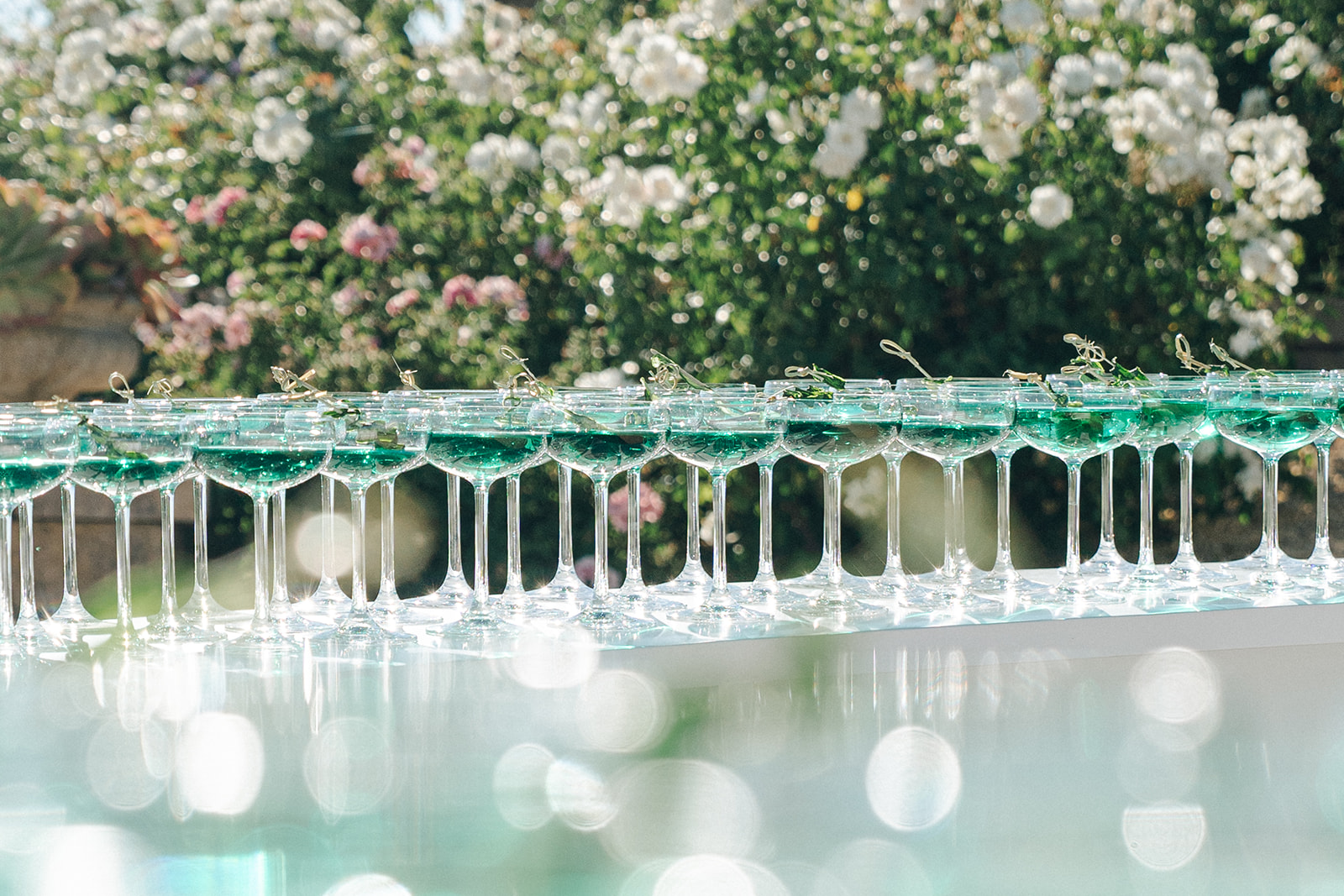  A line of elegantly arranged champagne glasses filled with a vibrant green drink, set against a lush garden with pink flowers in the background.