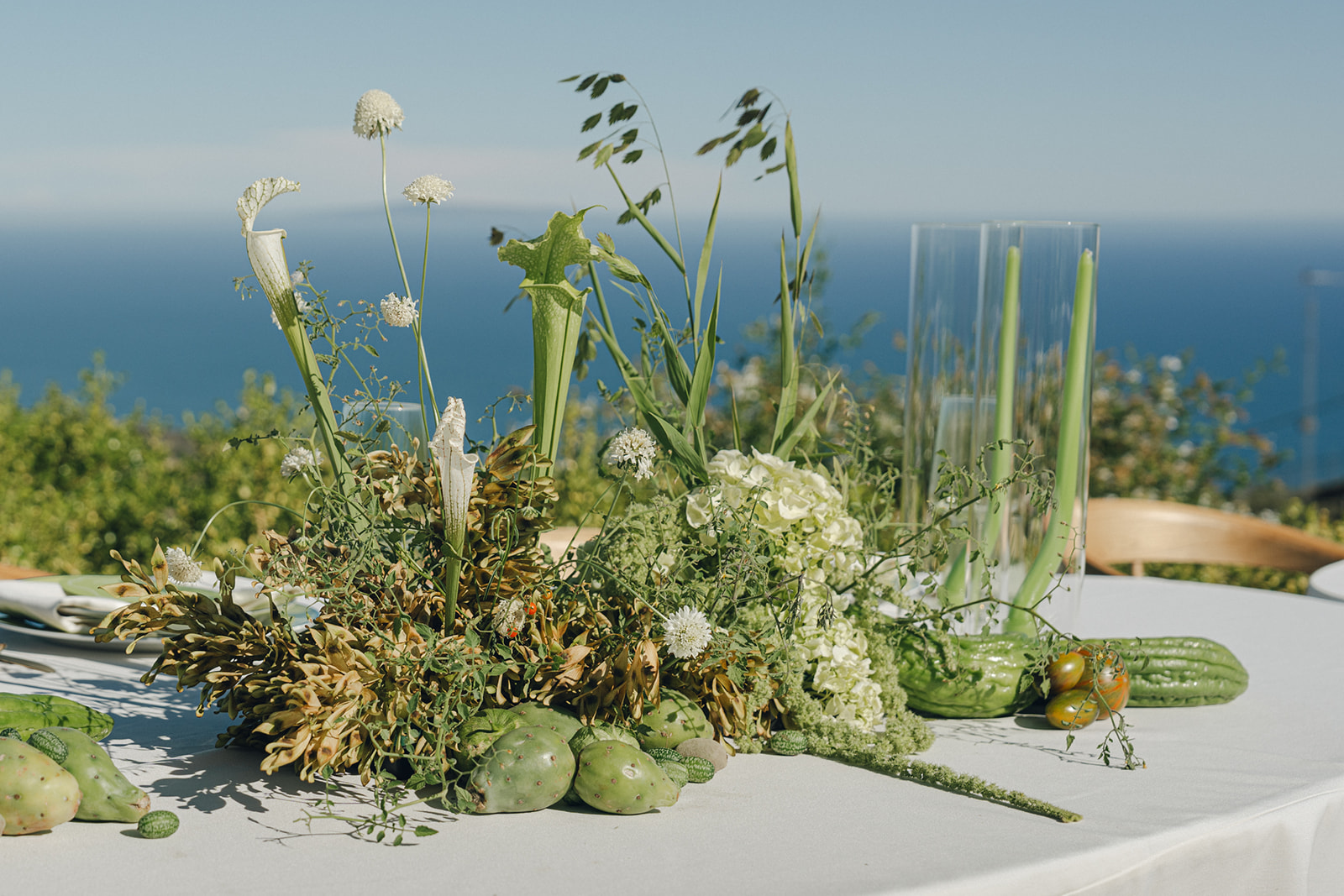A beautiful wedding reception table, with a stunning floral centerpiece featuring a variety of green plants, succulents, and wildflowers, set against a clear blue sky and ocean view.