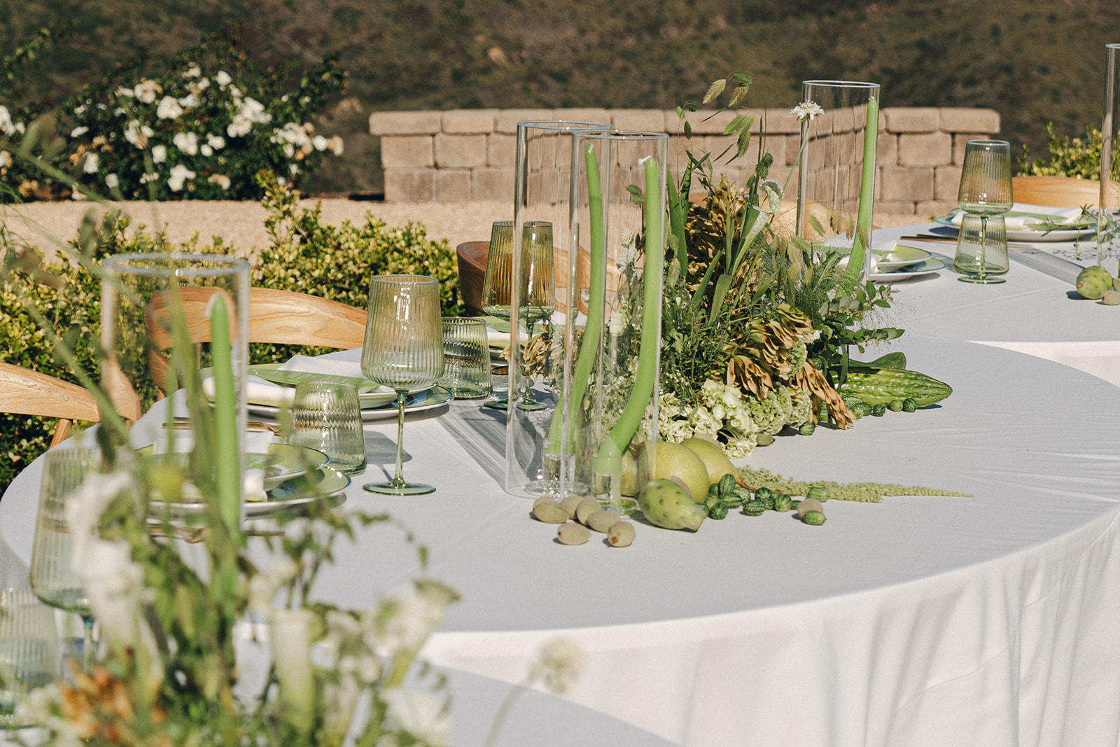 A beautifully set outdoor dining table with soft green tones. The table is adorned with tall glass vases containing flowers, green apples, and delicate foliage, creating a fresh, elegant look with a scenic backdrop.