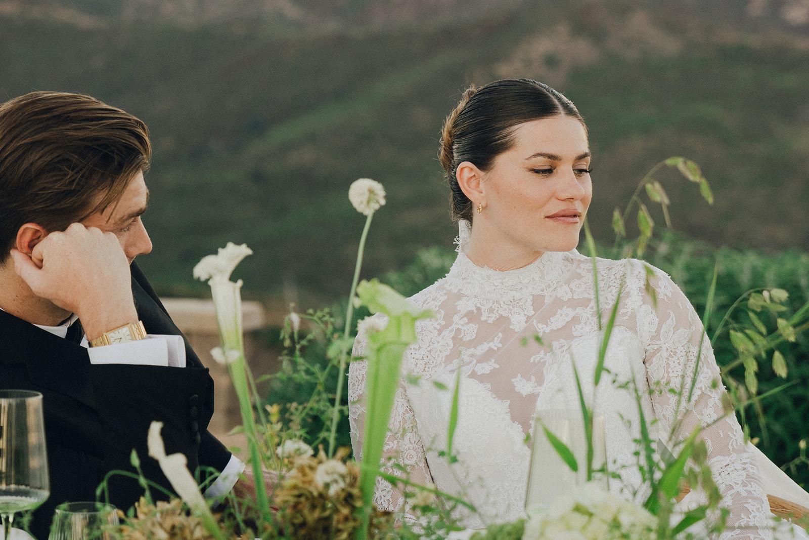 A candid moment of a bride in a lace wedding dress sitting at a table, with her partner beside her and floral arrangements surrounding them.