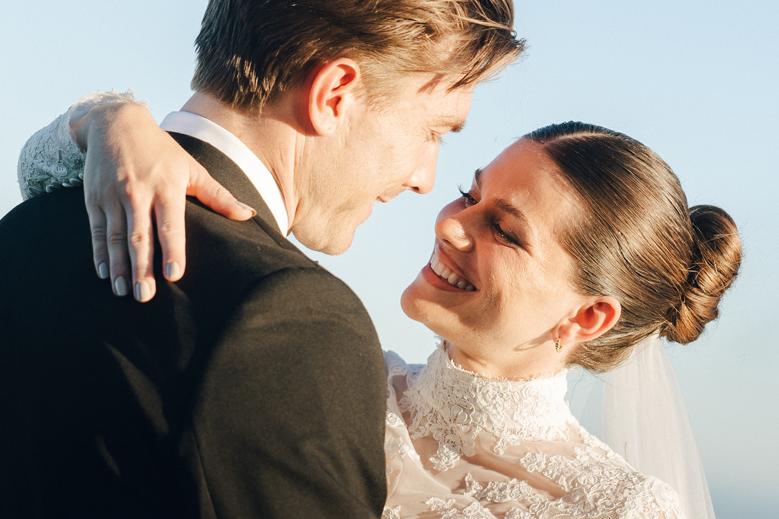 A joyful bride in a high-neck lace wedding dress smiles warmly at her groom, who is dressed in a classic black suit, as they share a loving moment outdoors.