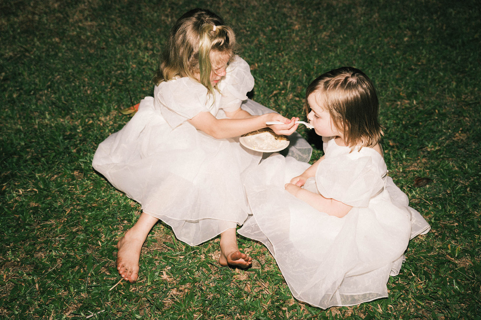 Two young girls in white dresses sitting on the grass, one feeding the other with a spoon, enjoying a moment together.