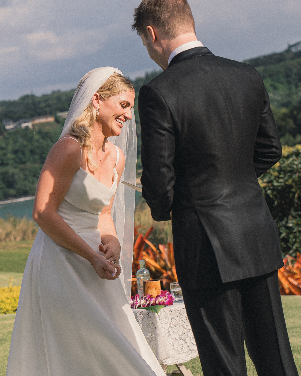 A bride laughing as she interacts with her groom during an outdoor wedding ceremony with lush greenery and a lake in the background.