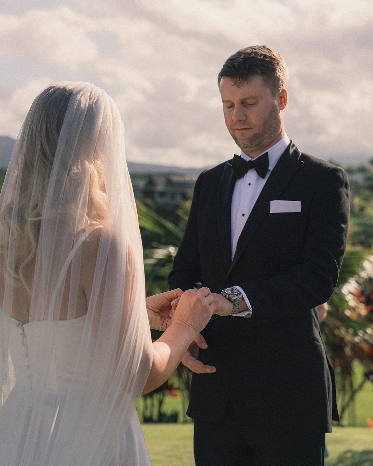 A bride placing a watch on the groom’s wrist during their outdoor wedding ceremony, with a backdrop of tropical greenery.