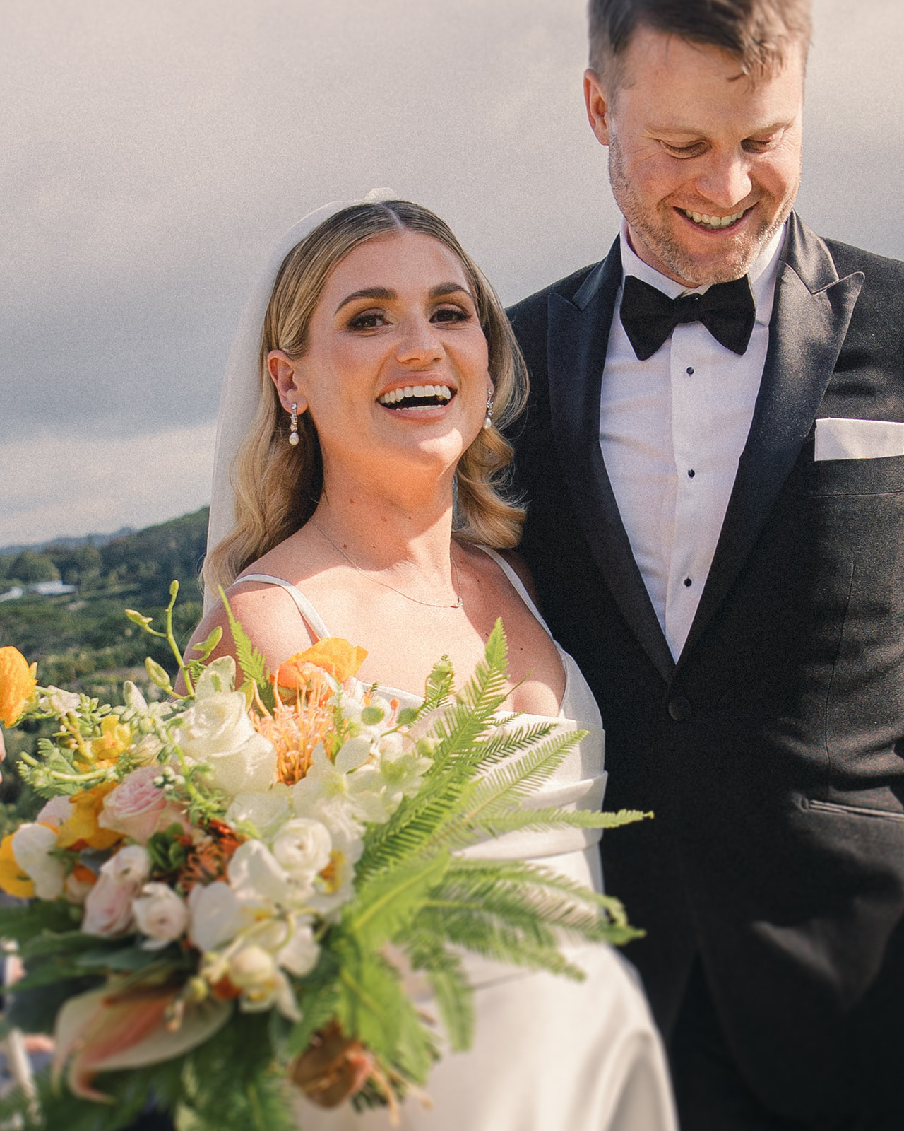 A bride holding a vibrant bouquet of orange and white flowers, smiling joyfully beside her groom in a tuxedo.