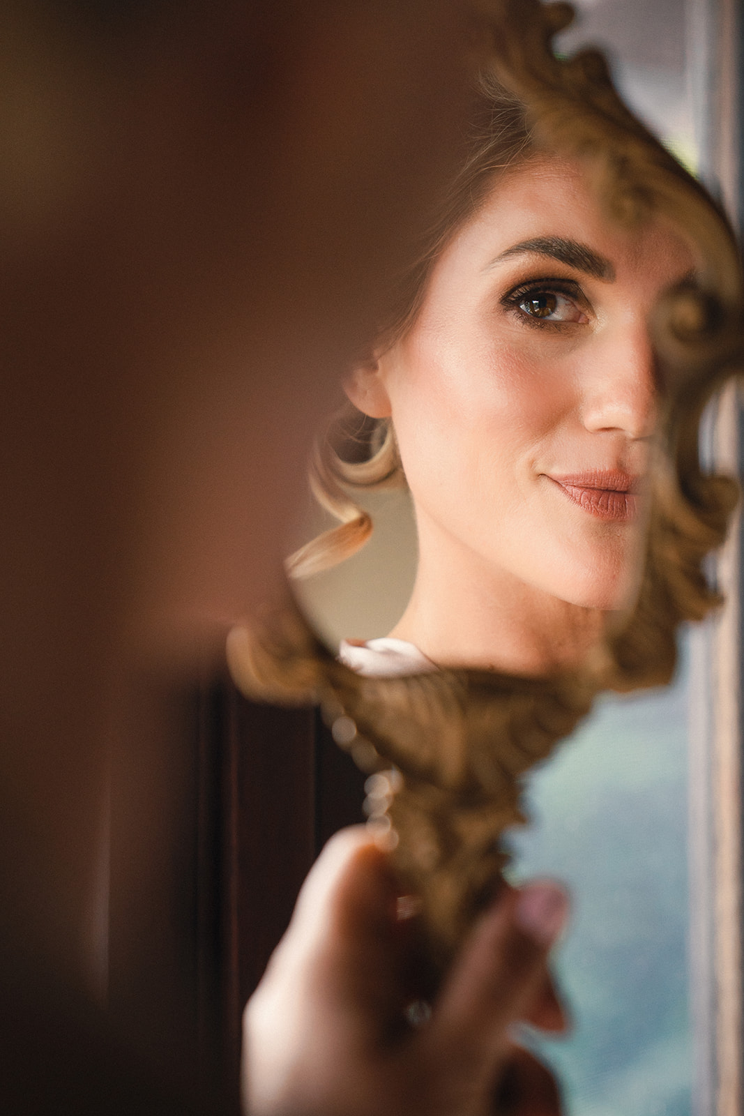 A close-up of a bride looking into an intricately framed mirror, capturing her soft smile and elegant makeup. The blurred foreground adds depth, highlighting her poised and graceful expression.