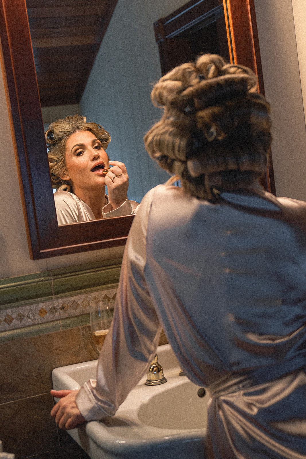 A bride is seen applying lipstick while wearing a silk robe, her hair in rollers, as she prepares for the big day. The mirror reflection captures her focused expression in a warm, softly lit space.