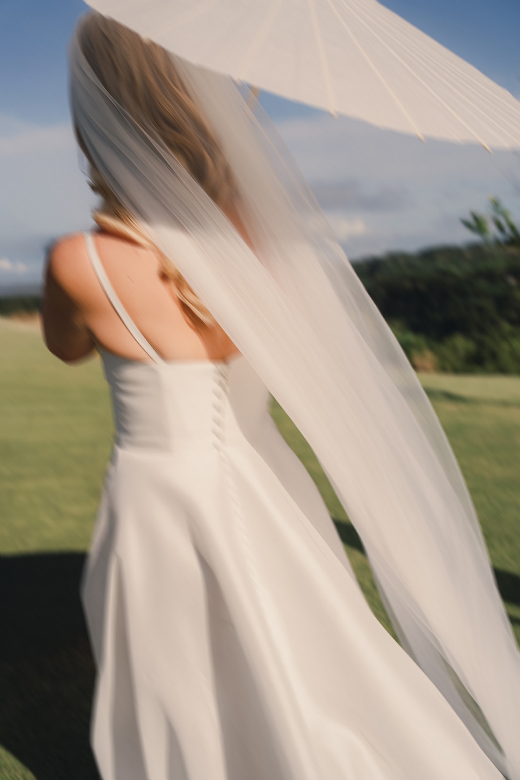 A bride is elegantly walking outdoors, her white gown flowing behind her as she holds a delicate parasol. The soft breeze lifts her veil, creating a dreamy, romantic feel against the lush green landscape.