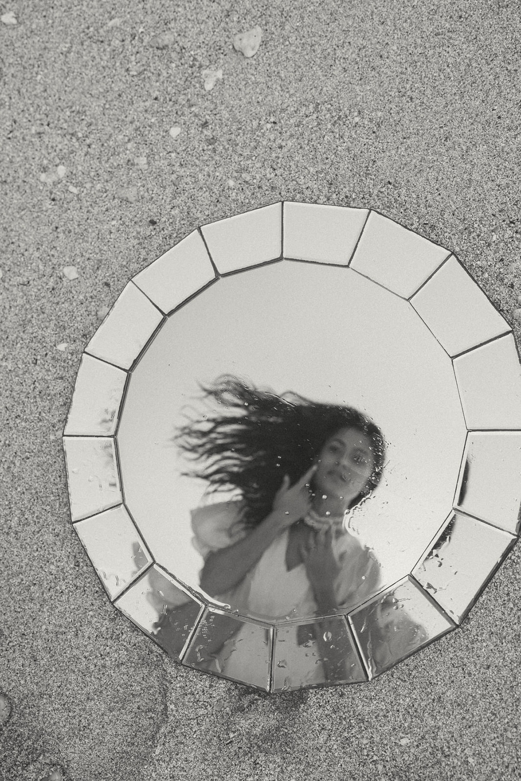 A black-and-white photo of a woman reflected in a round mirror on the sand. She is touching her neck and looking at the camera with her hair flowing in the breeze.