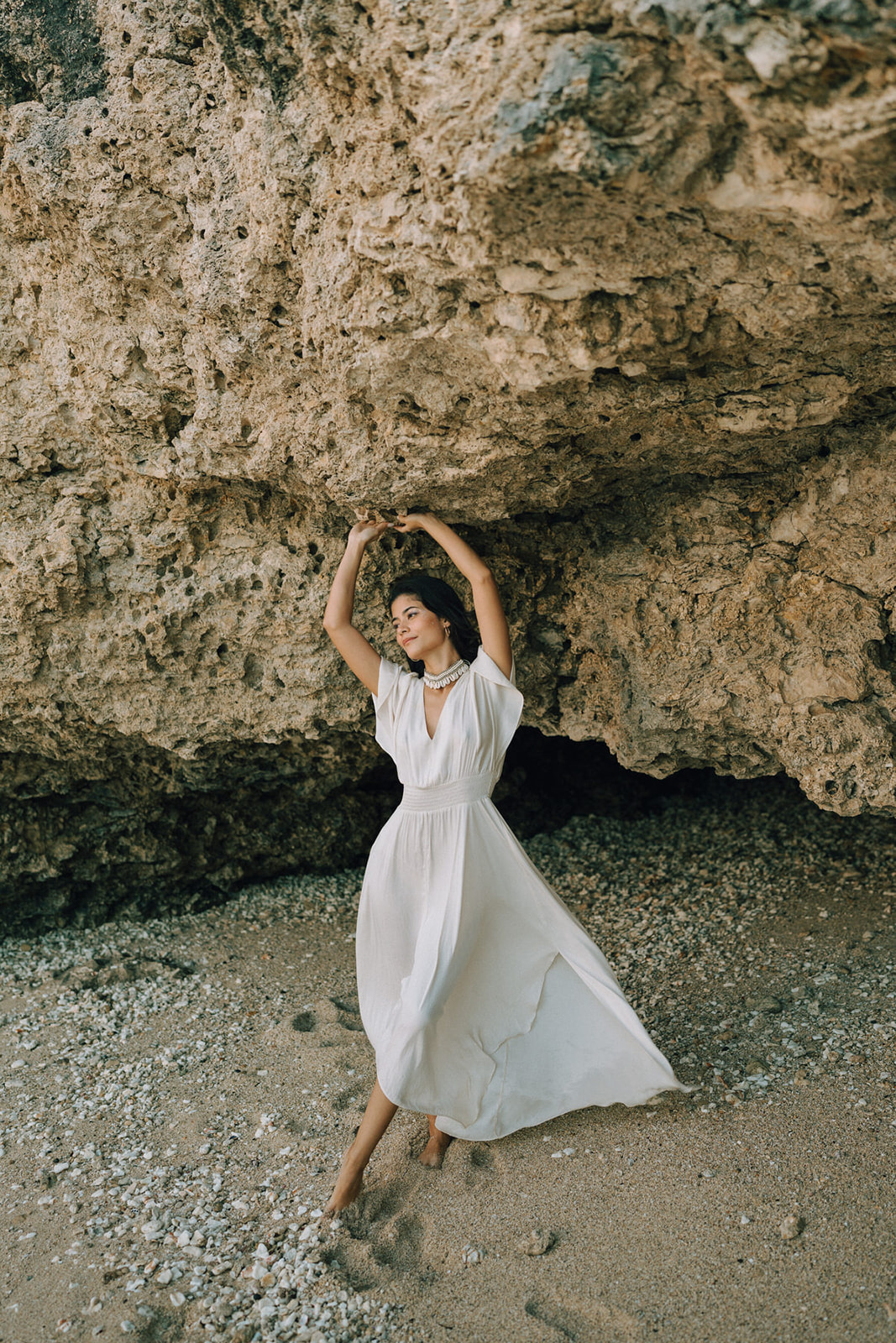 A woman in a flowing white dress, striking a dramatic pose while holding onto a rock near the shore, surrounded by a rugged coastal backdrop.