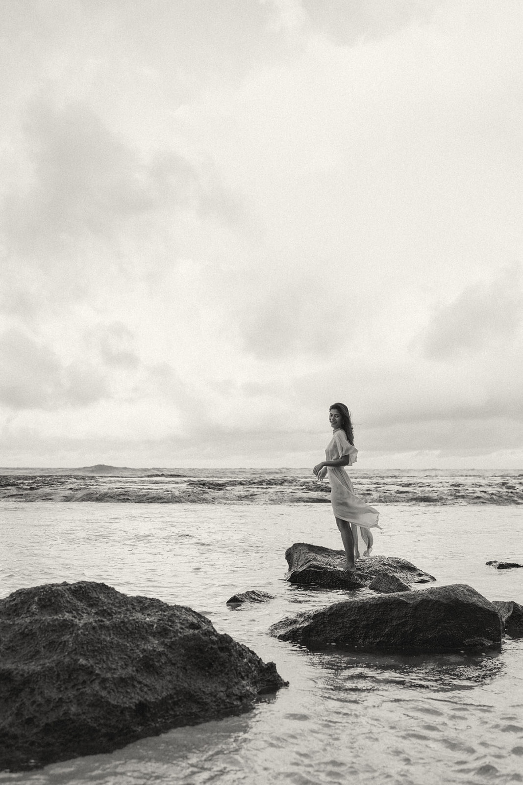 A woman standing on rocks in the water, with a wide ocean view and cloudy skies, capturing a peaceful and natural moment.