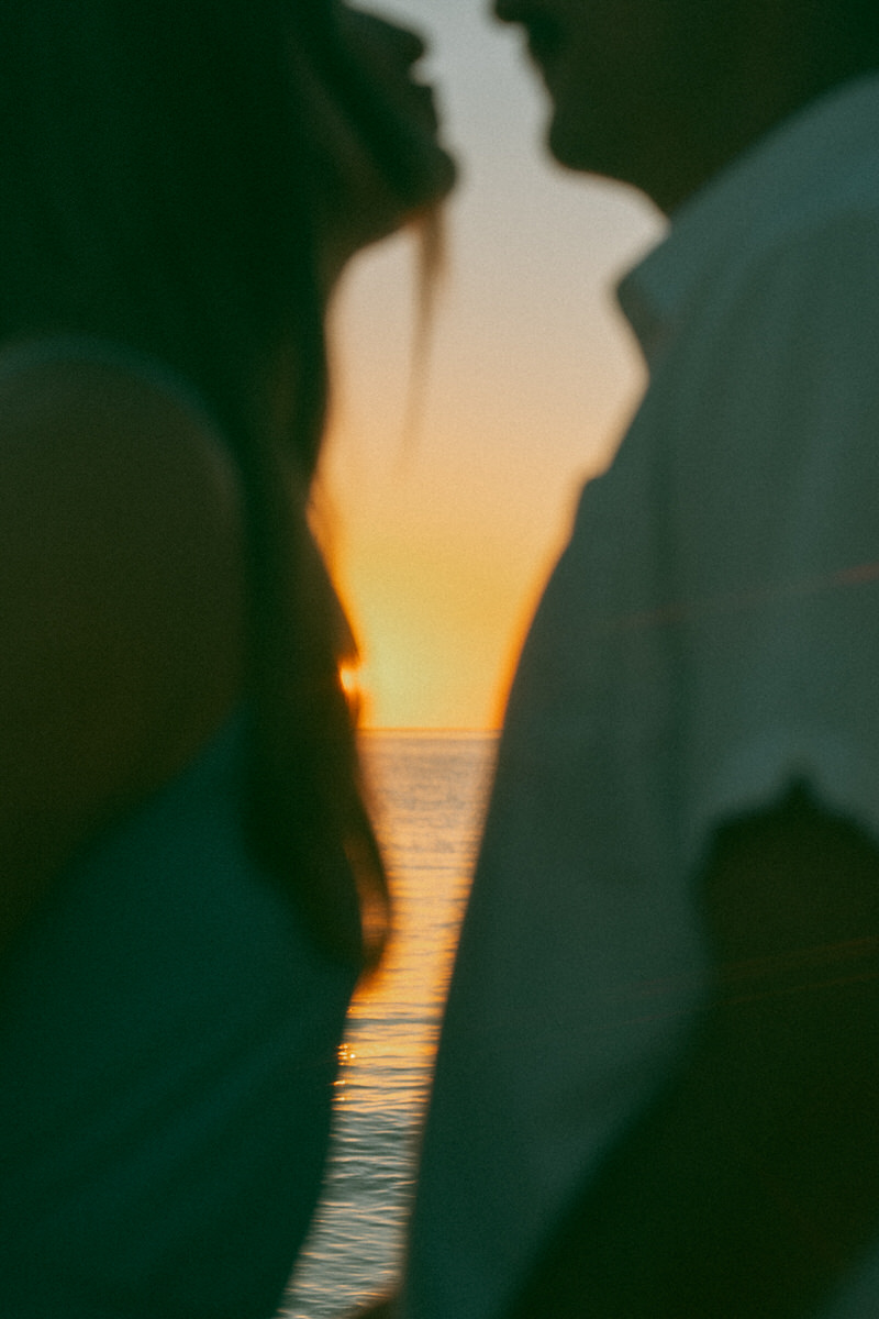 A soft-focus image of a couple sharing a kiss at sunset, with the vibrant orange hues of the ocean and sky providing a romantic backdrop. The image captures their closeness from a side profile.